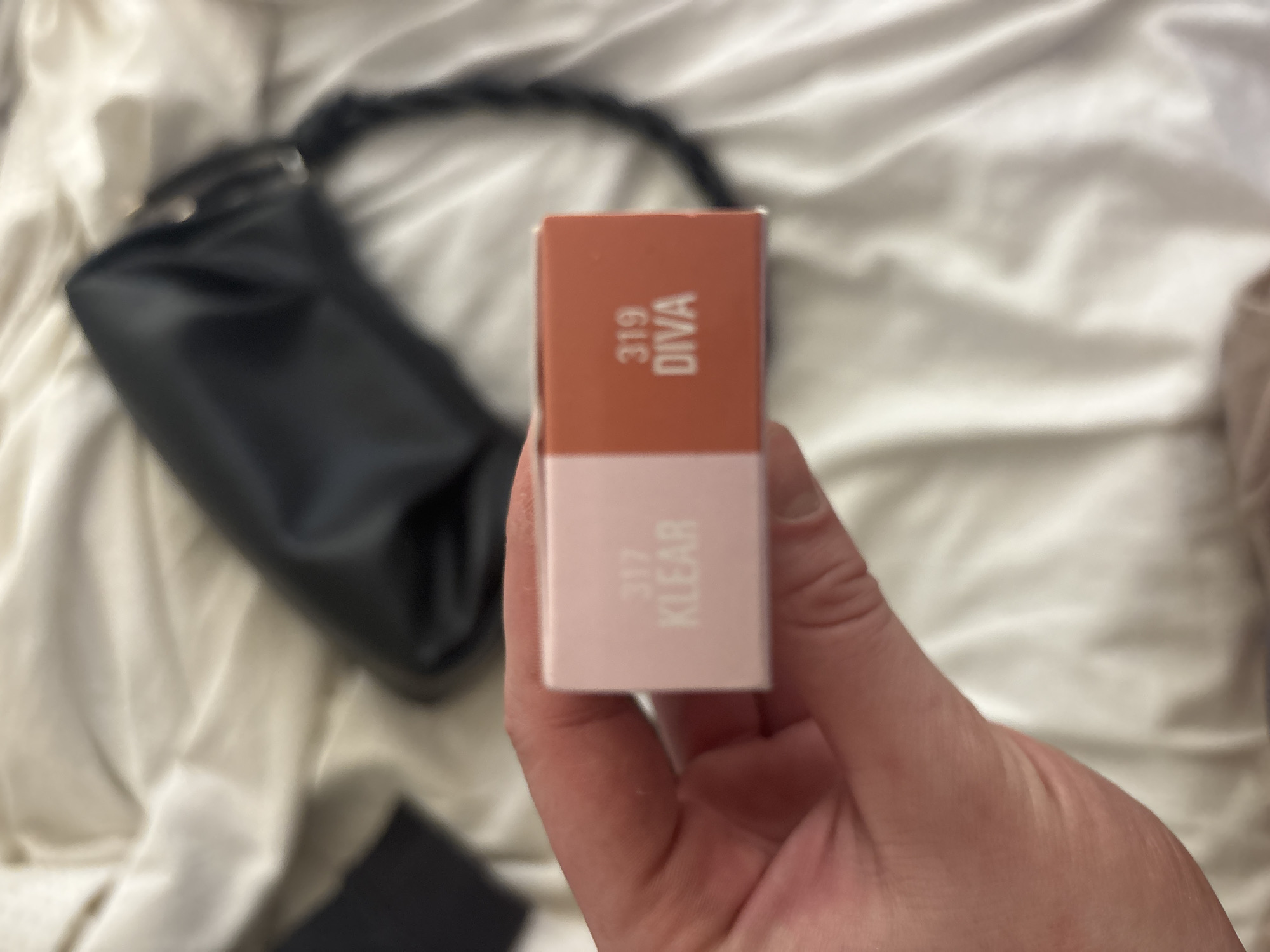 A close-up shot features a light-skinned hand holding a small, rectangular cosmetic product box. The box is divided into two colored sections: the top is a peachy-orange with the white, vertically oriented text "319 DIVA," and the bottom is light pink with the white, vertically oriented text "317 KLEAR." This suggests two different shades or product types.
The background is softly blurred, showing a dark, possibly black, bag or purse with a braided strap positioned on the left side. The bag rests on a light-colored, textured fabric, likely a bedsheet or blanket, which appears to be cream or off-white and has gentle folds. The scene is set indoors, possibly in a home in Buenos Aires, Argentina, under soft, diffuse lighting. The focus is sharp on the product box, making its details clearly visible, while the background elements provide context without distracting from the main subject.