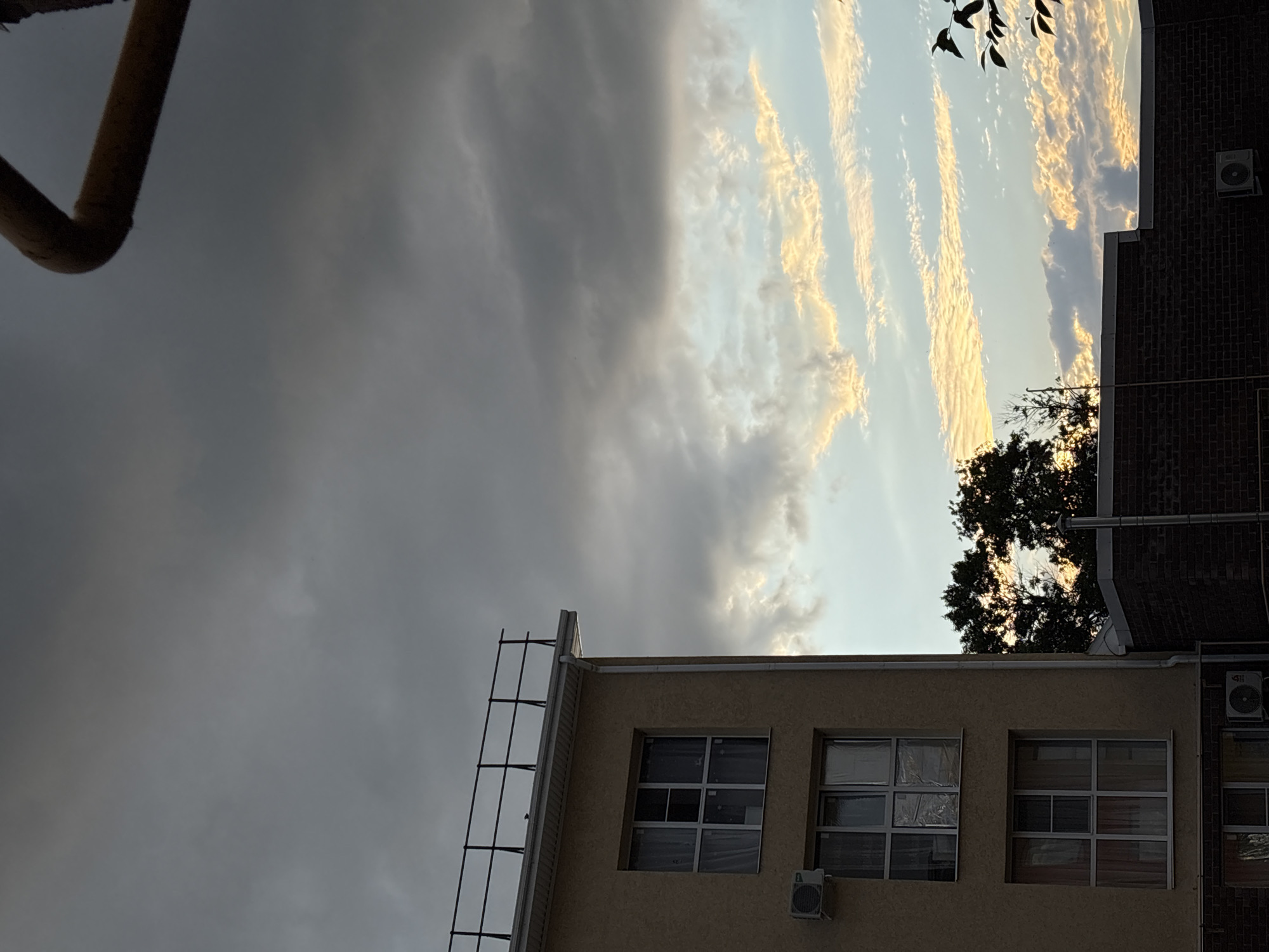 The image is a low-angle shot of buildings against a dramatic sky. The sky is a mixture of dark, ominous clouds on the left and a brighter expanse on the right, filled with wispy, golden-tinged clouds, suggesting either sunrise or sunset. A portion of a rusty metal pipe is visible in the upper left corner.
In the foreground, the top of a light-colored building is shown with several windows. The windows are paneled, with some covered in protective sheeting. An external air conditioning unit is mounted below one of the windows. To the right, a darker brick building rises, also featuring an air conditioning unit and a drainpipe. A metal ladder-like structure is attached to the side of the light-colored building.
Silhouetted against the bright sky are the branches of a tree, with a few leaves visible. The overall scene conveys a sense of an urban or semi-urban environment, possibly residential or commercial, during a transitional period of the day. The weather appears to be partly cloudy, with distinct changes in light and cloud cover. The mood is somewhat dramatic due to the sky's appearance. No text is visible in the image.