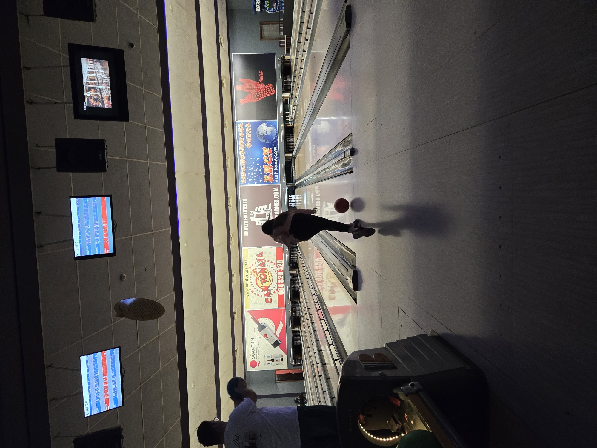 The image shows a bowling alley. A person is in the process of bowling, releasing the ball down the lane. Nearby, two people are standing by the bowling equipment. Advertisements, including one for "CANTONATA 064 820 320" which is located in Pleven, Bulgaria, are placed above the lanes. Electronic scoreboards are visible on the wall.