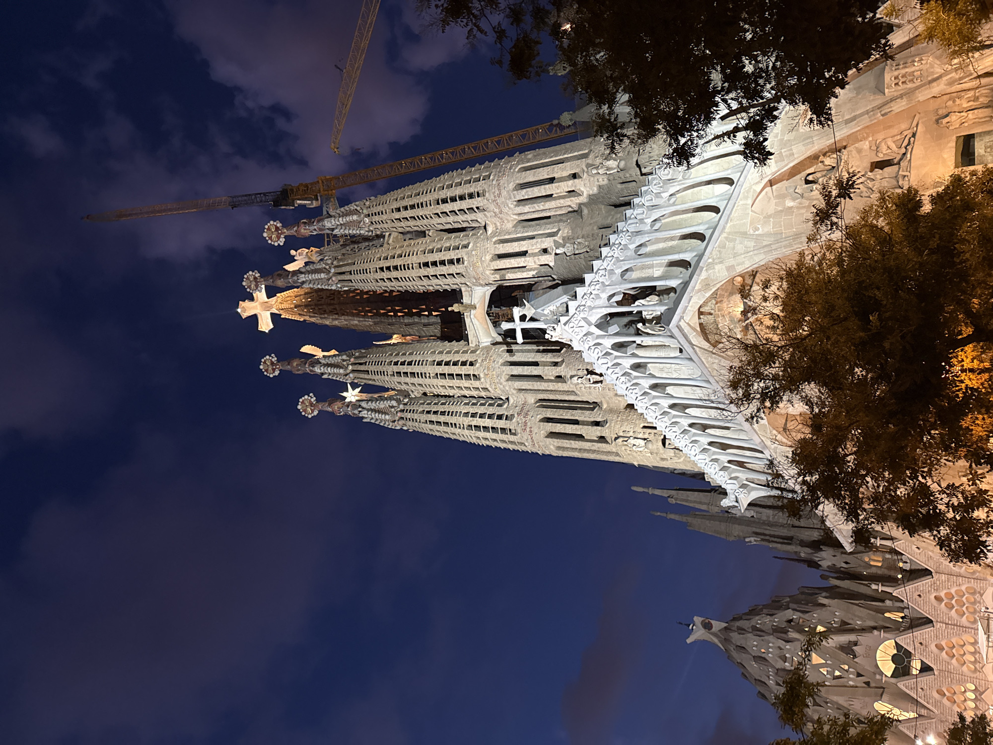 The image shows a low-angle, nighttime view of the Sagrada Familia in Barcelona, Spain. The iconic basilica's spires, adorned with intricate details and illuminated from within, dominate the frame. A large construction crane is visible to the right of the central spires, with another smaller crane in the background. The sky above is a deep blue, with scattered, wispy clouds that are slightly illuminated. Trees with dark foliage are in the foreground, partially obscuring the lower sections of the basilica's facade. The overall scene captures the monumental architecture of the Sagrada Familia against a twilight sky, with evidence of ongoing construction.