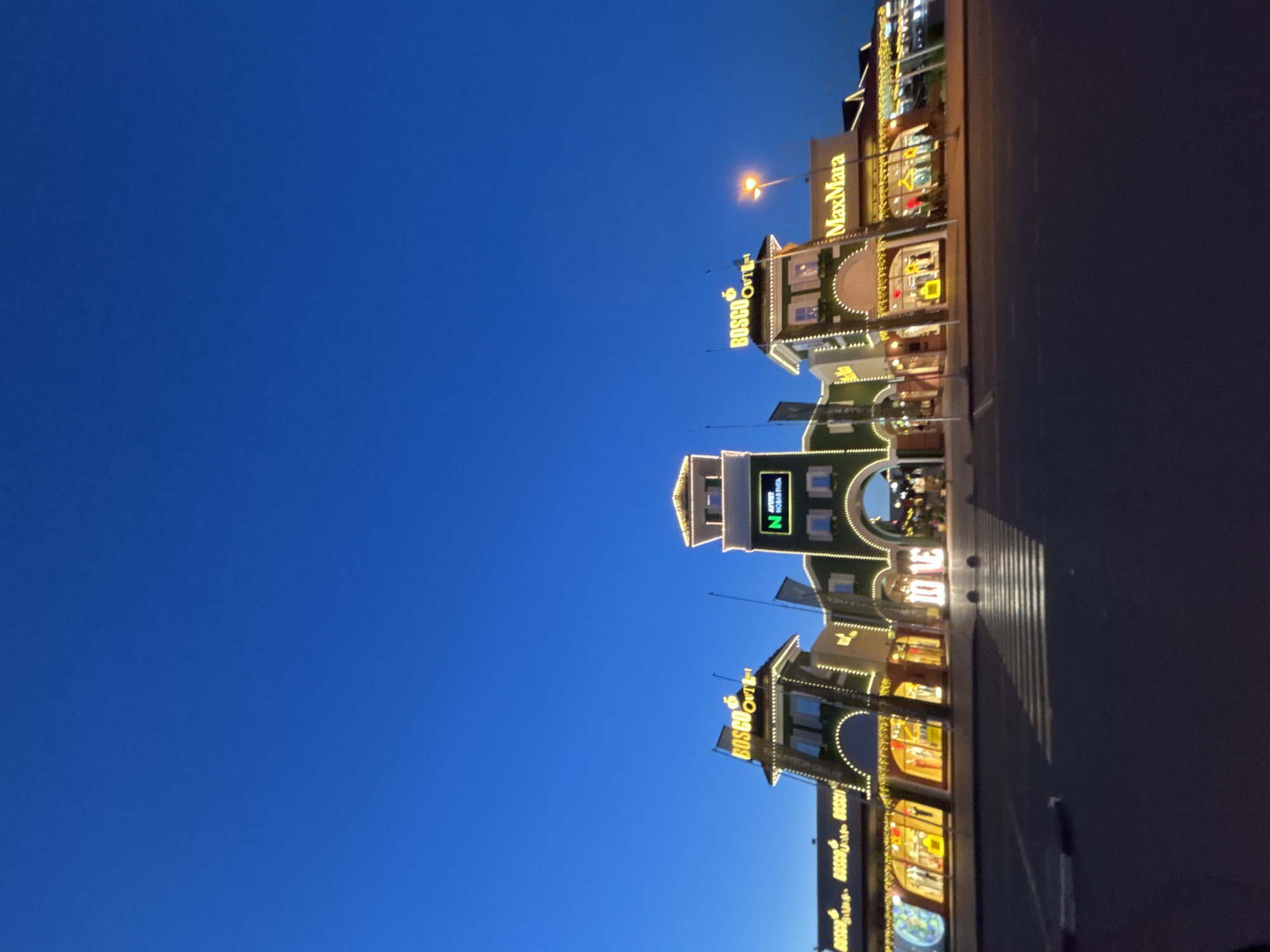 The image shows a facade of a shopping center at dusk in Pokrovskoe, Russia. The architecture is in a classical style with arched entrances and decorative lighting. Signs for "BOSCO Outlet," "NAVERT HOBAЯ PHAA," and "MaxMara" are visible, indicating the presence of branded stores. The shopping center is illuminated with numerous yellow lights, creating a festive atmosphere against the dark blue twilight sky. In the foreground, a pedestrian crossing with white stripes is marked on the dark asphalt. The scene is empty of people, suggesting it is either early in the evening or the area is not heavily frequented at this time. The overall impression is a well-lit commercial area designed for shopping.