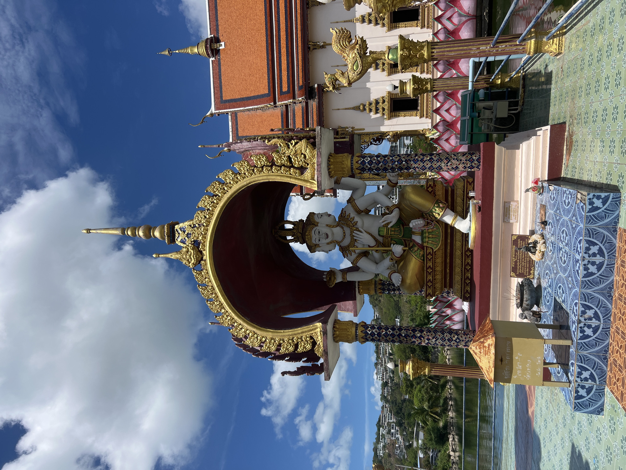 The image depicts a religious monument in Baan Plai Laem, Thailand. The monument features a four-faced statue of a deity, possibly Brahma, positioned within a large, ornate archway. The statue is light-colored with gold accents. The archway is red inside and decorated with gold ornamentation. The surrounding architecture includes a red-roofed building with gold embellishments and supporting pillars decorated with lotus patterns. The ground is tiled. In the background, a body of water and foliage are visible, along with a sparse settlement. The sky is blue with scattered clouds.