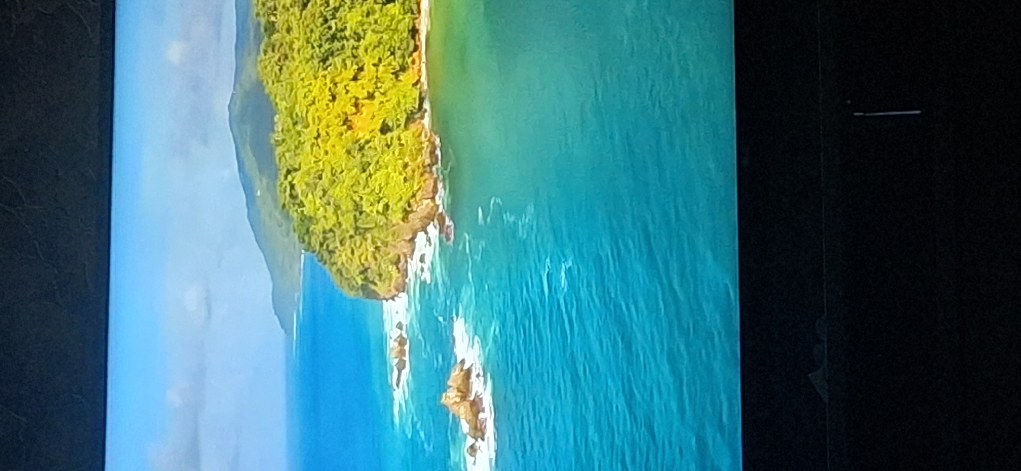 The image shows a tropical coastline with lush green vegetation covering a cliff face that juts out into the turquoise ocean. Several rocky outcrops are visible in the water, with waves crashing against them. In the background, a mountain range covered in similar vegetation stretches along the horizon under a clear blue sky with scattered clouds. The bottom portion of the image is black, suggesting it is a photograph taken of a screen. The location in Bila Tserkva, Ukraine, is not identifiable from the provided image, as the scene depicts a tropical environment, not characteristic of Ukraine.