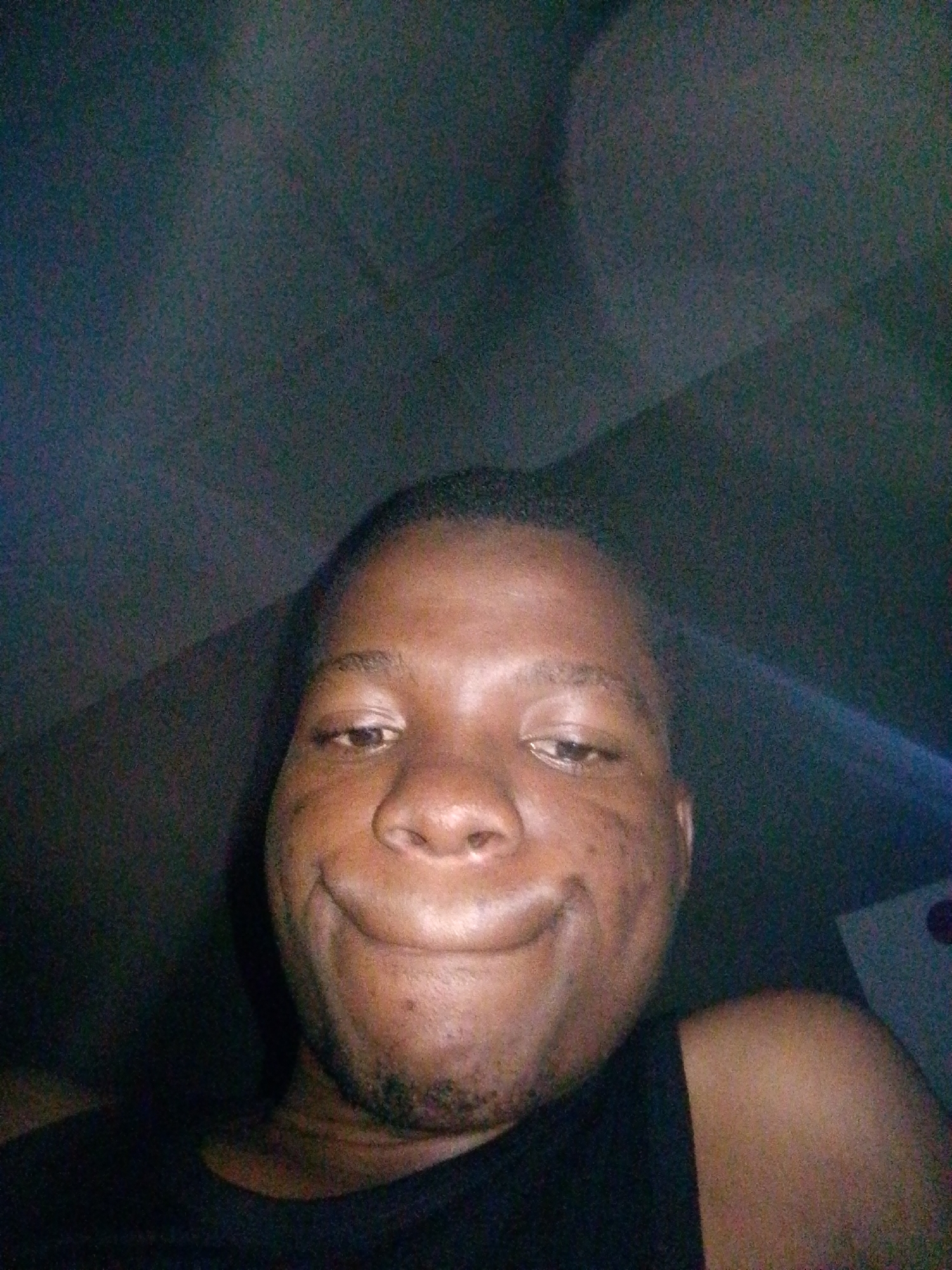 The image is a close-up, low-resolution selfie of a Black male, appearing to be in his late teens or early twenties, with short hair and a dark complexion. He is wearing a black sleeveless top and has a broad smile, showing teeth. His head is tilted slightly upwards, and his face is turned towards the camera. The background is dark and appears to be an interior space, possibly a car or a dimly lit room, with some subtle texture suggesting a ceiling or upholstered surface. There is a faint, diffused light source, contributing to the overall grainy quality of the image. No other individuals or distinct objects are clearly visible. The setting does not provide specific visual cues to confirm a location within Ipetumodu, Nigeria.