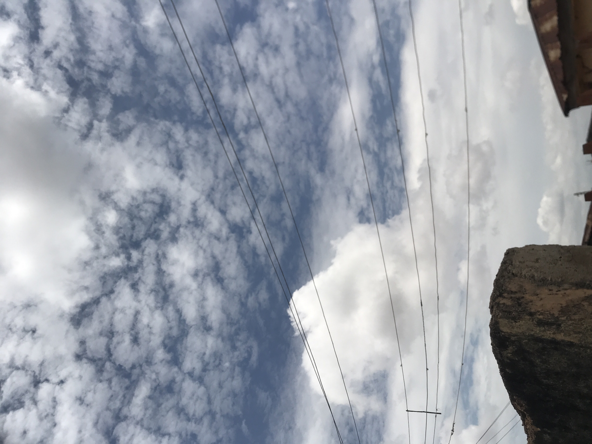 The image depicts an outdoor scene in Rafin Par, Nigeria, dominated by a partly cloudy sky with visible utility lines and fragments of structures at the bottom.
The upper two-thirds of the frame are occupied by a blue sky interspersed with various cloud formations. The top-left features numerous small, scattered white clouds, while a larger, dense white cumulus cloud is prominent in the mid-left. The upper-right shows a bright, possibly overexposed, expanse of white cloud.
Approximately five parallel, dark utility lines, likely electrical or communication cables, traverse the mid-section of the image horizontally. A short, dark, vertical line connects to one of these horizontal lines on the far left. Several minute, dark, indistinct specks are observable along these cables.
In the bottom-left quadrant, a dark, rectangular, roughly textured structure, consistent with weathered concrete or stone, is partially visible. Its upper surface is darker than its side, which shows lighter, reddish-brown tones. The bottom-right quadrant reveals the upper part of a building, identifiable by a corrugated, weathered reddish-brown roof, indicative of a metal material.
No human figures, animals, or explicit actions are present in the image.