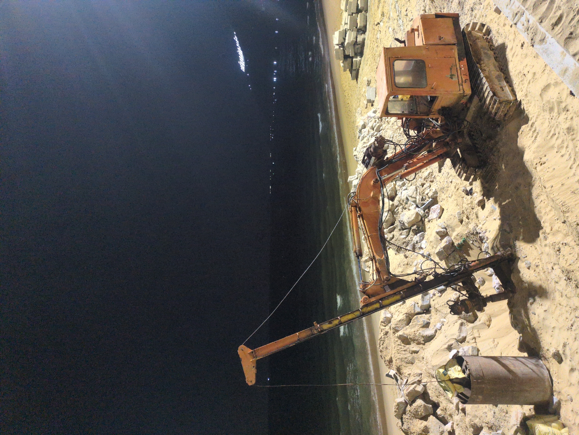 The scene depicts a piece of heavy construction equipment, an excavator, on a beach in South Quy Nhon Ward, Vietnam, at night. The excavator is positioned on a sandy area near the shoreline. A stack of square concrete blocks is visible adjacent to the excavator. The ocean is visible in the background, with scattered lights across the horizon. A large cylindrical pipe lies in the sand near the excavator's boom.