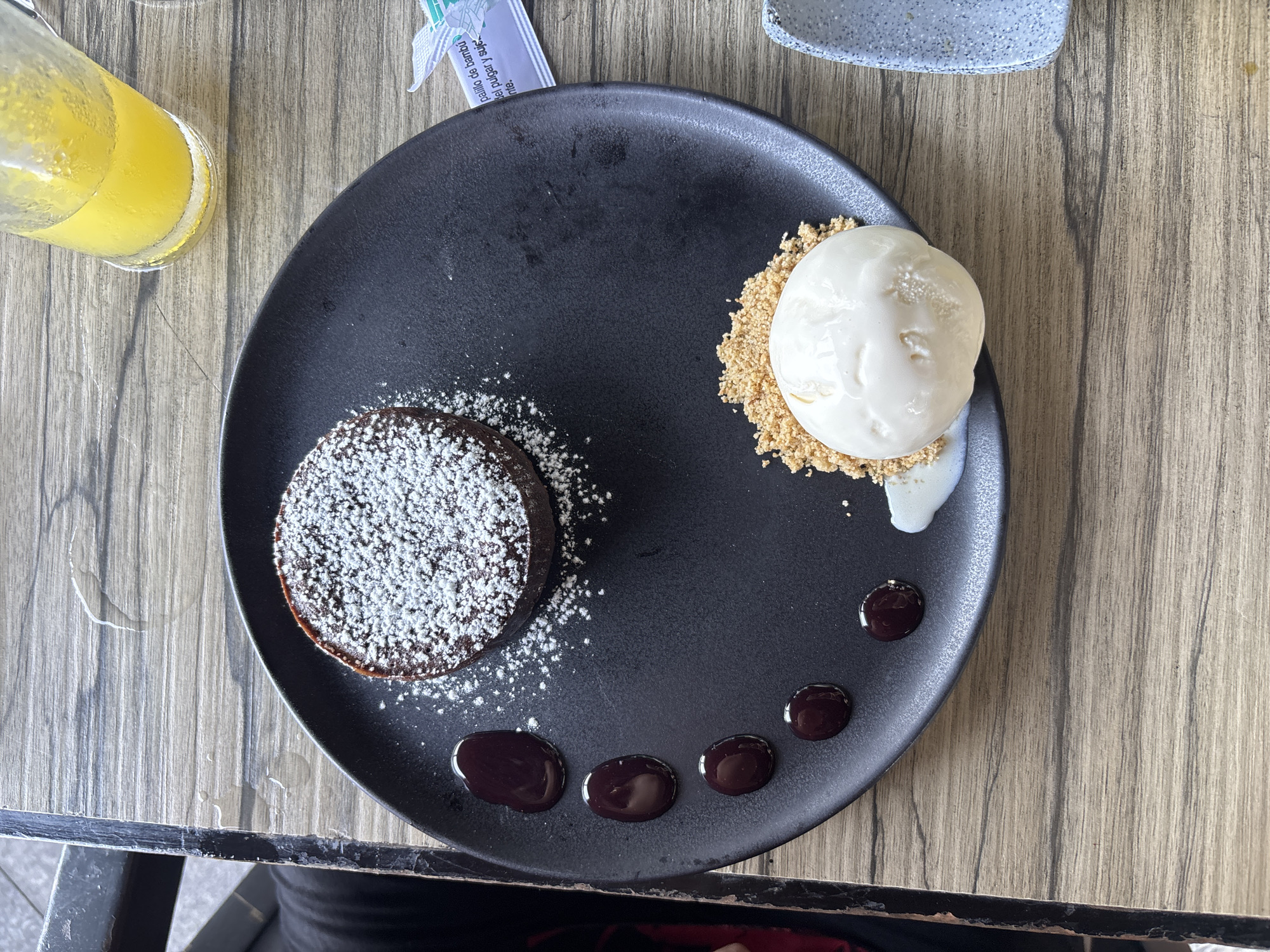 A top-down view of a black plate on a wooden table shows a chocolate lava cake topped with powdered sugar alongside a scoop of vanilla ice cream on a bed of crushed graham crackers, and five small droplets of chocolate sauce arranged in a semi-circle. A glass of orange juice sits to the left, and a sugar packet is visible at the top. Based on the image details, it is not possible to determine the specific location within Veracruz, Mexico.