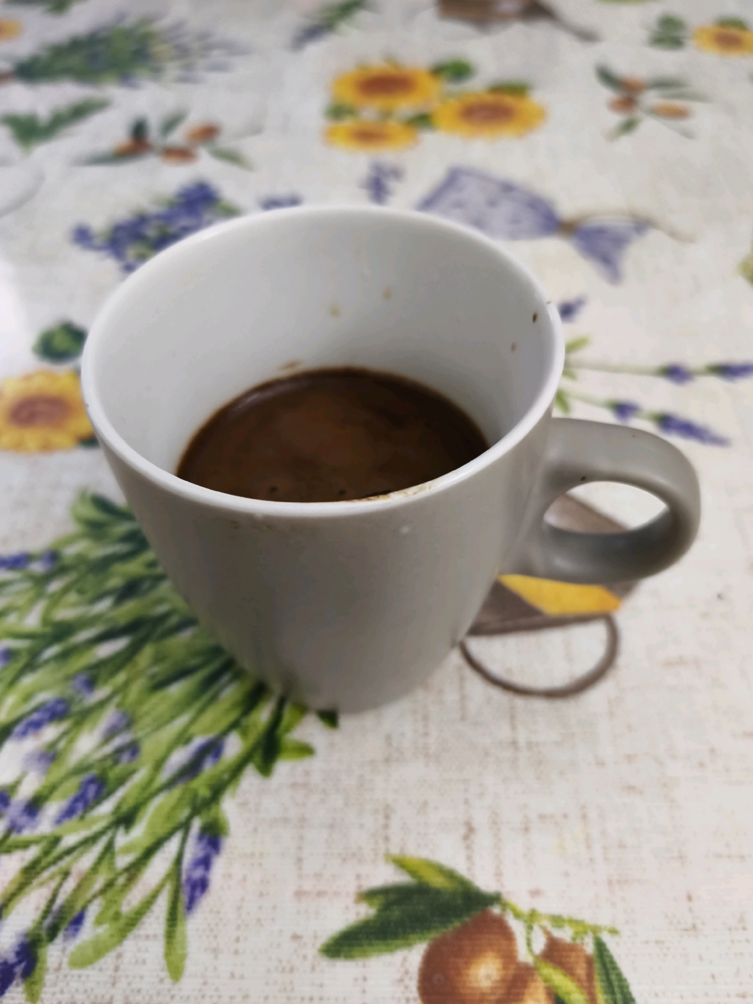A close-up, slightly blurry, high-angle shot shows a gray mug filled with dark coffee sitting on a tablecloth with a floral and fruit pattern. The mug is positioned to the right of center, with its handle clearly visible and facing right. The coffee within the mug has a foamy surface. The tablecloth features a repeating pattern of lavender sprigs, sunflowers, and what appear to be lemons or small oranges. The background is an interior setting, likely a table in a home. The image does not contain any identifiable features that would place it in Livorno, Italy, or any specific city.