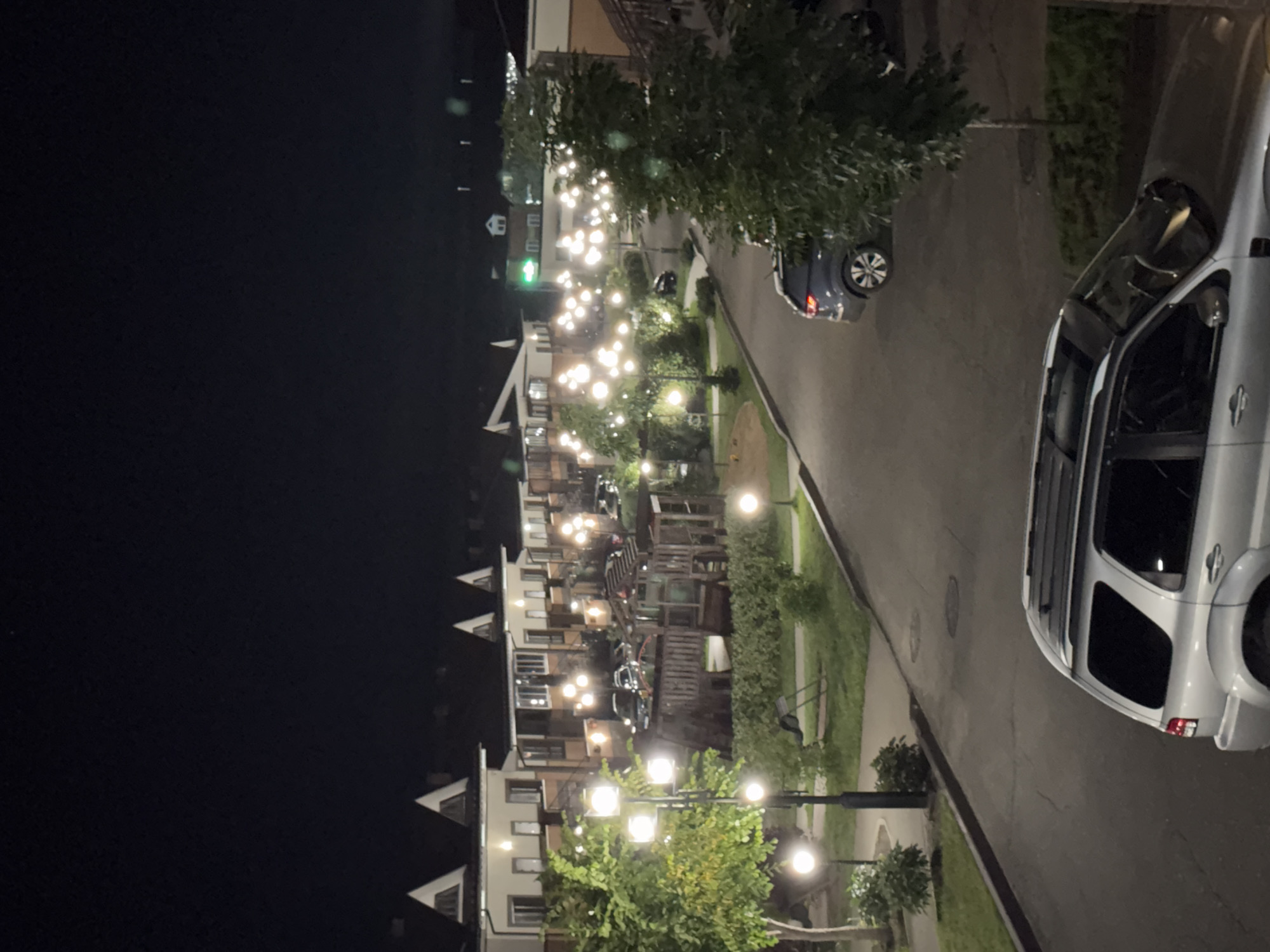 The image shows a night scene in a residential area, possibly a courtyard, in Markova, Russia. The foreground features the rear section of a silver SUV parked on a paved surface. The middle ground reveals a pathway lined with lampposts casting warm light on trees and a playground with wooden structures. Several cars are parked along the pathway. In the background, a row of multi-story houses with illuminated windows and pointed roofs are visible. The sky is dark, indicating nighttime.