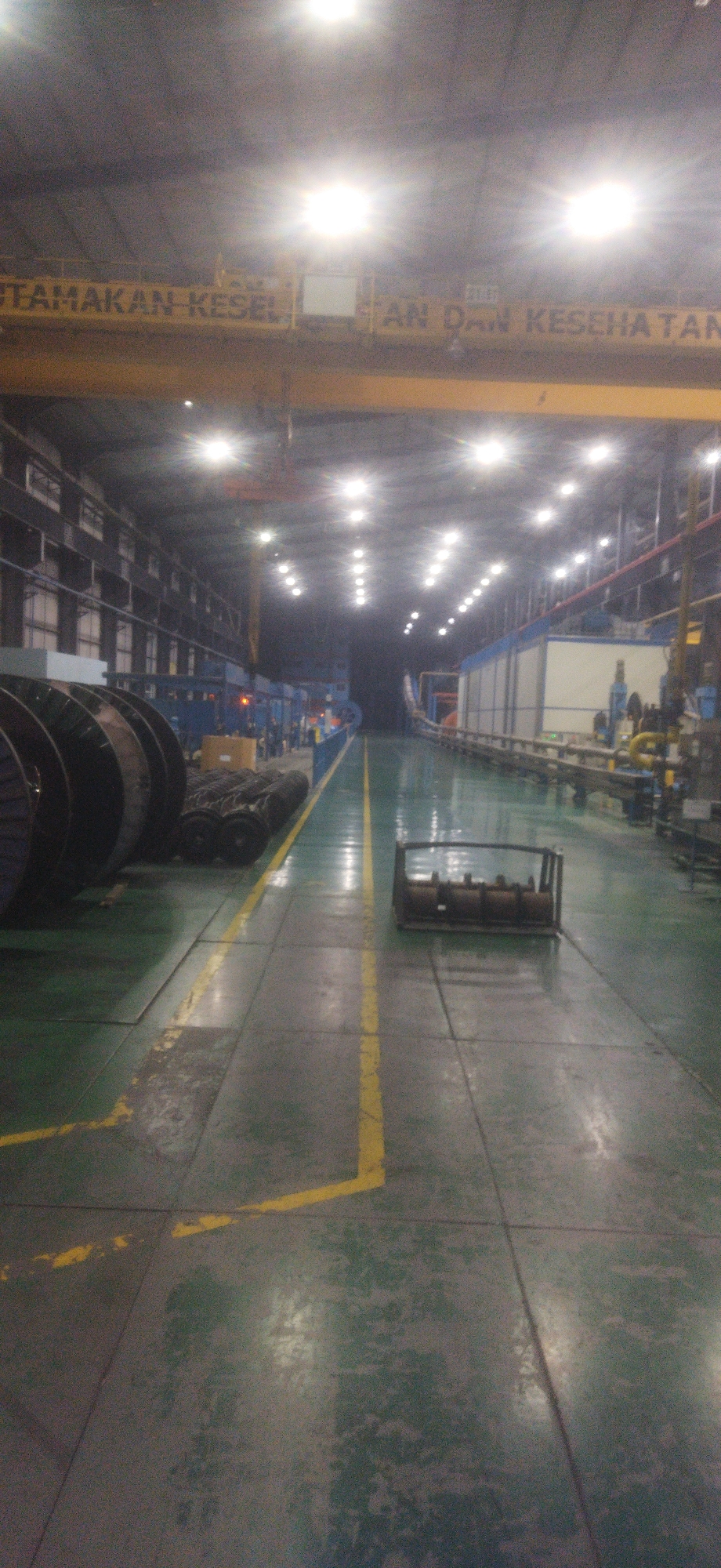 An interior view of a large factory floor in Cileungsi, Indonesia, shows a long, well-lit corridor. The floor is concrete, painted green, with yellow lines marking pathways. Large, dark-colored spools of cable are stacked on the left. Further down the corridor, several people are visible, appearing to be workers, engaged in various tasks. The factory is equipped with industrial lighting, and the environment appears to be a production or manufacturing facility. A yellow sign spanning the width of the upper portion of the image has text that is partially legible, reading "UTAMAKAN KESEL..." and "...AN DAN KESEHATAN". This likely translates to "Prioritize Safety..." and "...and Health," a common slogan in Indonesian workplaces.