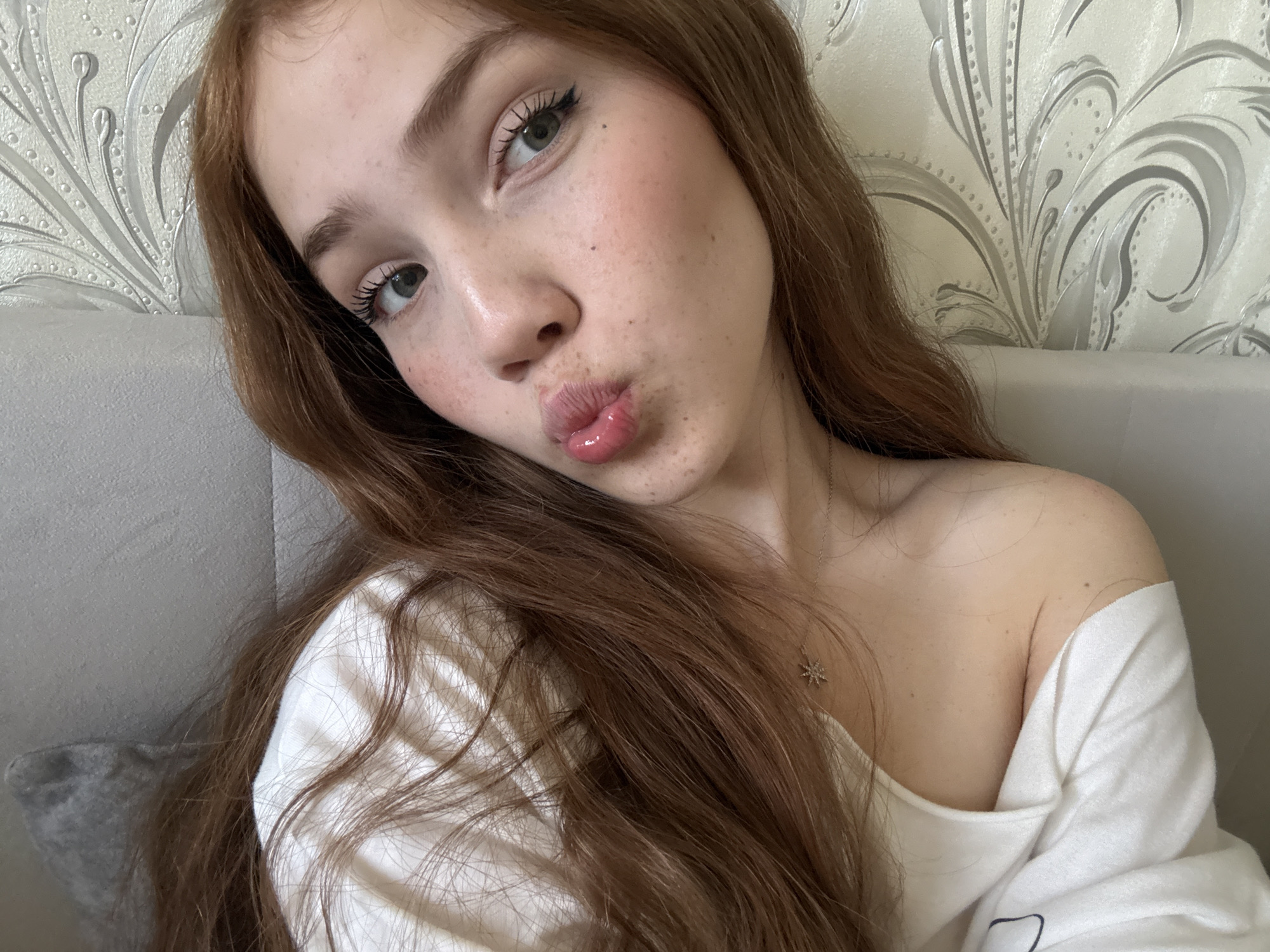 A close-up, eye-level selfie shows a young woman with long, wavy reddish-brown hair cascading around her. She has a light complexion, green eyes with winged eyeliner and visible mascara, and a scattering of freckles across her nose and cheeks. Her lips are pursed in a "pouty" or "kissing" expression, enhanced with a glossy, light pink tint.
She is wearing a white, off-the-shoulder top, revealing her left shoulder and collarbone. A delicate necklace with a small, sparkling, star-shaped pendant is visible around her neck.
The background features a light-colored wallpaper with an ornate pattern of grey and white swirling floral or leaf motifs. To the left and behind her, a solid light grey headboard or cushion is visible. The lighting is soft and even, suggesting an indoor setting. The overall mood of the image is casual and playful. There is no discernible text within the image.