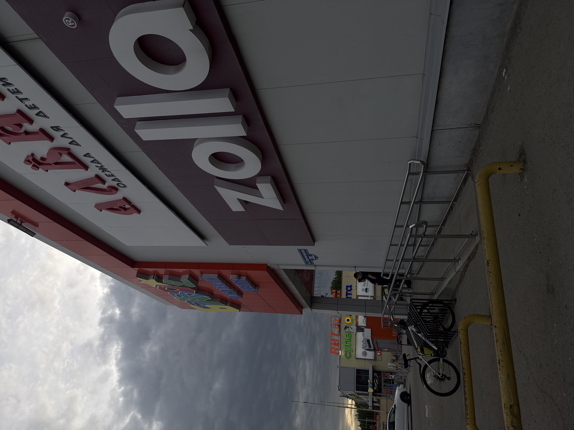 The image shows an exterior view of the "Zolla" clothing store at a shopping center in Perm, Russia. The store's sign is visible, along with a smaller sign indicating "Clothing for Children". The sky is cloudy. A bicycle is parked near a metal railing on the sidewalk. Other store signs are visible in the distance, indicating a shopping area. A person wearing a mask is standing by the entrance.