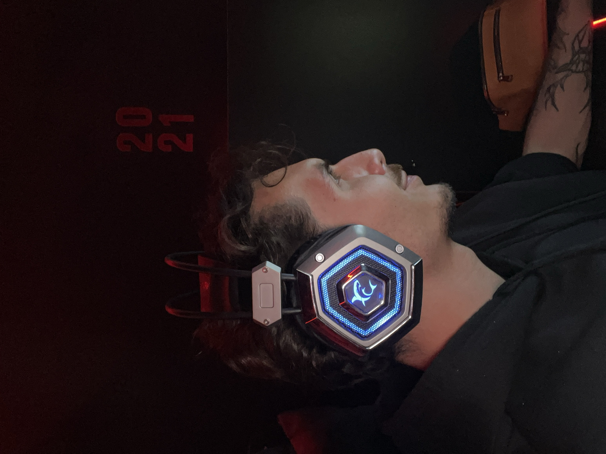 The image displays a young man lying down in a dark, dimly lit environment, viewed in profile facing right. He wears elaborate over-ear gaming headphones featuring hexagonal earcups with silver-grey trim and striking blue LED lighting. A central, glowing blue shark logo is prominent on the visible earcup. The man has dark, curly hair, a mustache, and a short beard or heavy stubble. His expression appears neutral and relaxed, with his eyes seemingly closed or looking upwards. He is dressed in a black hoodie or shirt.
The background is predominantly dark, almost black. To the left, subtle red illumination highlights the area, where red numbers "20" and "21" are visible, oriented sideways. In the upper right portion of the image, a light brown fanny pack or small bag with a zipper is visible, likely hanging. Below it, a tattooed arm, featuring dark, possibly abstract or tribal designs, is partially shown. The overall setting suggests a private, leisure space, likely indoors and at night or in a room with controlled lighting, in Budva, Montenegro. The man's posture and headphones indicate he is engaged in an activity such as gaming or listening to audio.