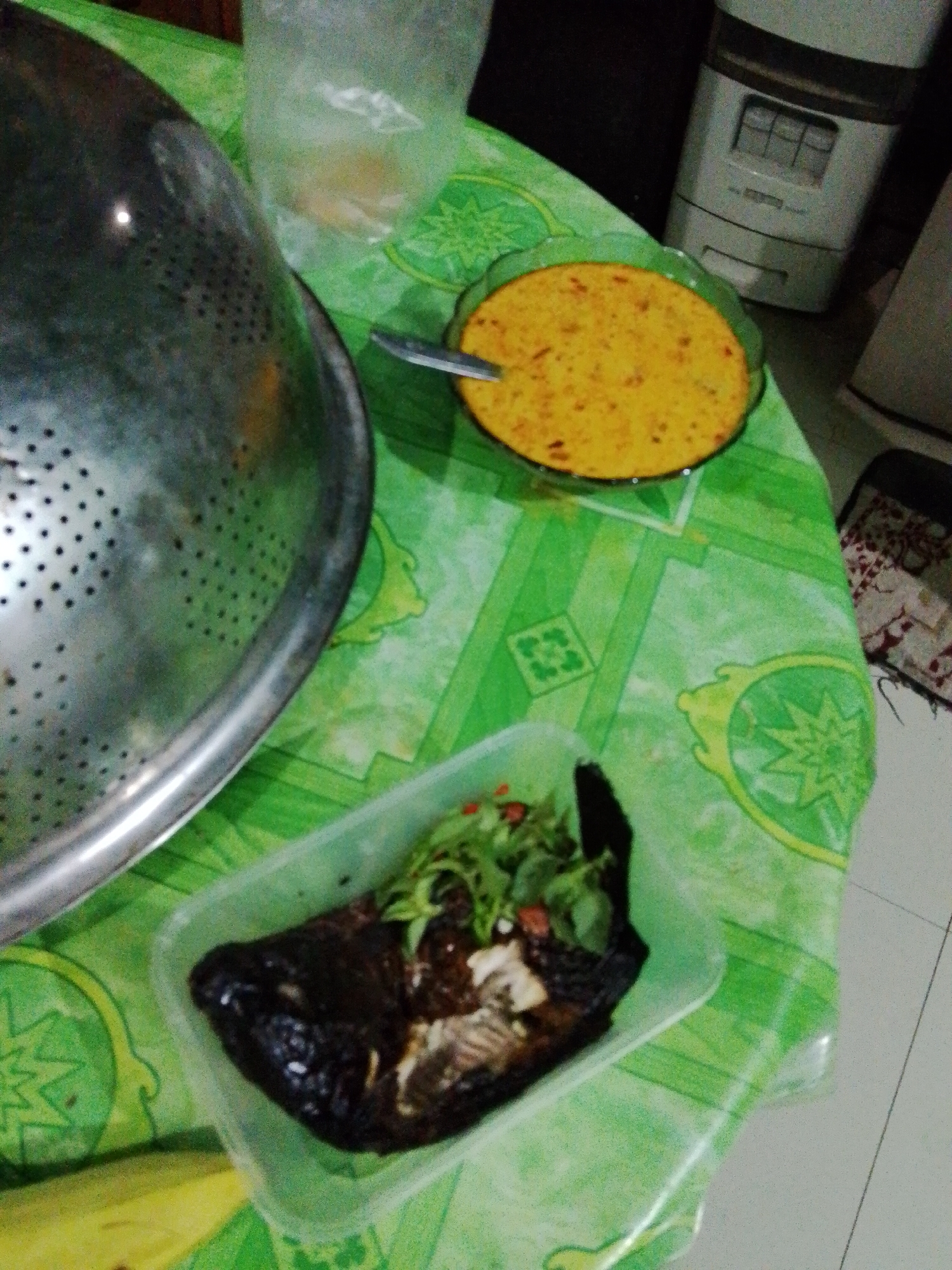 A high-angle shot captures a meal laid out on a green tablecloth patterned with yellow and green floral motifs. In the foreground, a rectangular plastic container holds a dark-colored, grilled fish topped with chopped green herbs and red pieces of chili. To the right of the container is a round glass bowl filled with a vibrant orange curry. A silver spoon rests in the curry, with its handle extending out of the bowl. To the left of the frame, a large, metallic colander with numerous perforations is partially visible. In the background, a white kitchen appliance with a dark panel featuring small rectangular buttons is situated on a dark surface. The overall setting suggests a domestic kitchen or dining area in Sendangrejo, Indonesia, likely during meal preparation or consumption.