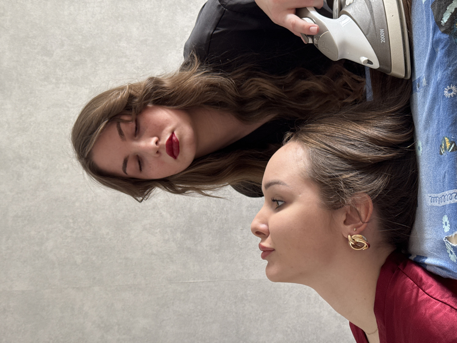 Two young women are depicted indoors in what appears to be a staged or unconventional photo. The image is oriented vertically, with subjects appearing to be lying down on a light-colored surface, possibly a floor or bed.
In the upper portion of the frame, one woman with long, light brown hair and red lipstick has her eyes closed. She is wearing a black top. A hand, appearing to be her own based on the matching black sleeve, holds a silver and white household iron. The iron, labeled "2000W," is pressed onto her own hair, which is spread out on the light surface. Her face shows freckles.
Below and to the right, a second woman is visible in profile, looking to the left. She has brown hair pulled back and is wearing a dark red top. She has a distinctive gold-colored, intertwined hoop earring. Her head rests on a blue fabric patterned with white flowers and small yellow butterflies, likely a pillow or bedding. Her eyes are open, and her lips are slightly parted.
The overall scene suggests an unusual activity where one woman is ironing her hair with a clothes iron, while another woman is casually lying nearby. The setting is simple, with a plain light background dominating the left side of the image. The event takes place in Chaikovsky, Russia.
