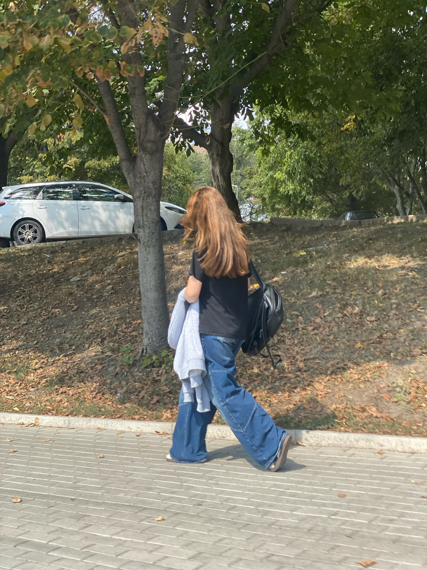 A person with long, auburn hair is walking along a paved sidewalk next to a grassy embankment. They are wearing a black t-shirt, loose-fitting blue denim jeans, and brown shoes. A grey hooded sweatshirt is draped over their left arm, and a black backpack is worn on their right shoulder. Behind the person, a white station wagon is parked on a paved area adjacent to the embankment. The embankment is covered in dry leaves and sparse green grass. Trees with green and yellowing leaves line the background, suggesting an autumn setting. The scene appears to be in an outdoor urban or park environment. The location is identified as being in the city of Chișinău, Moldova.