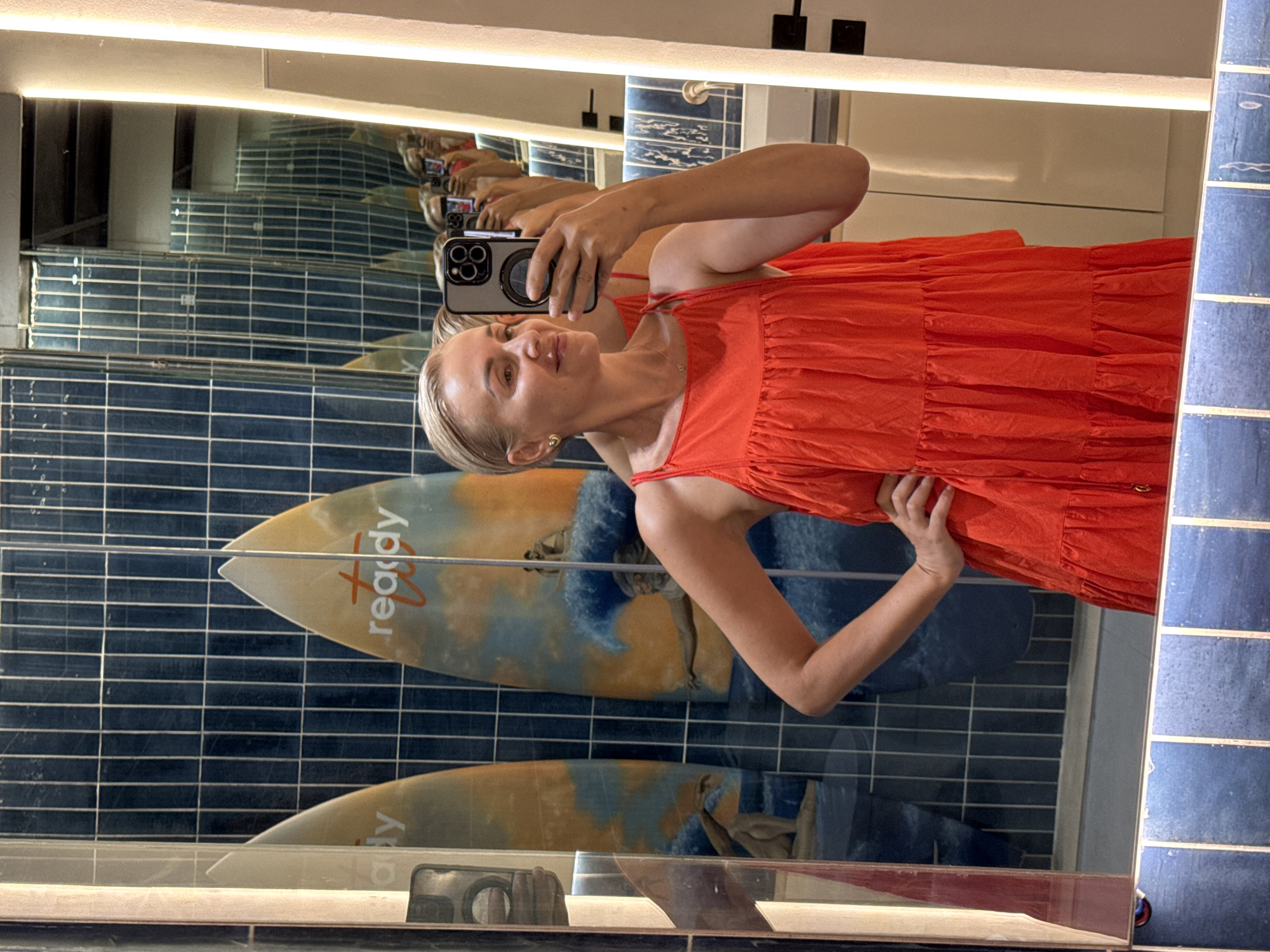 A woman takes a mirror selfie in a bathroom in Kuta Selatan, Indonesia. She has short blonde hair and wears a sleeveless, orange tiered dress. The bathroom wall behind her features blue tiled walls with two horizontal rows of surfboards with images of people surfing. The mirror reflects multiple images of the woman and the bathroom setup.