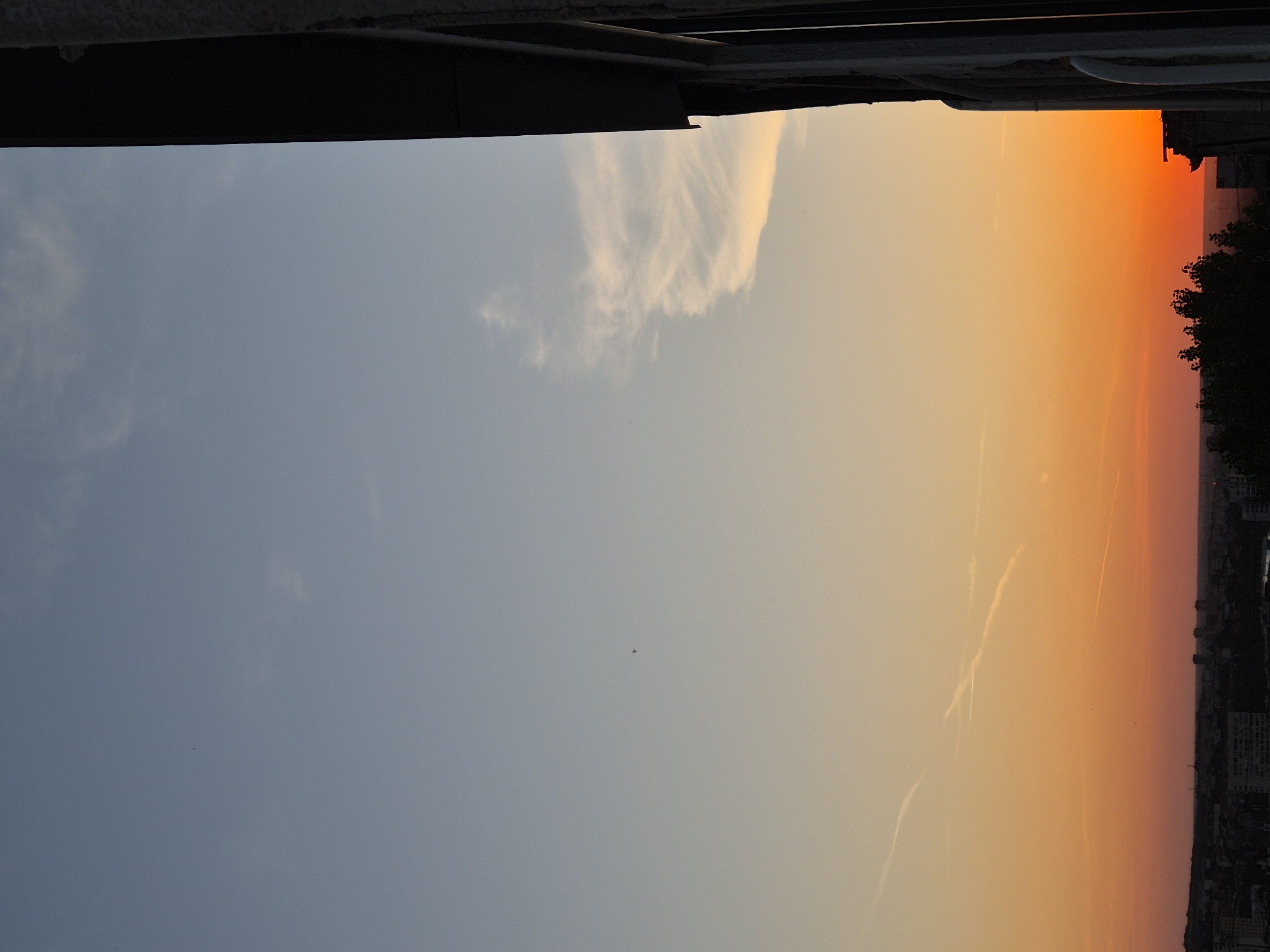 An outdoor shot captures a sunset over an unknown city. The sky transitions from a clear blue at the top to a vibrant orange and pink gradient near the horizon. Faint clouds are scattered across the sky, and contrails from airplanes are visible. In the foreground, the right side of the image is dominated by a dark, vertical structure, possibly part of a building, with a textured surface. Below the horizon, a cityscape with buildings and trees is silhouetted against the sunset.
