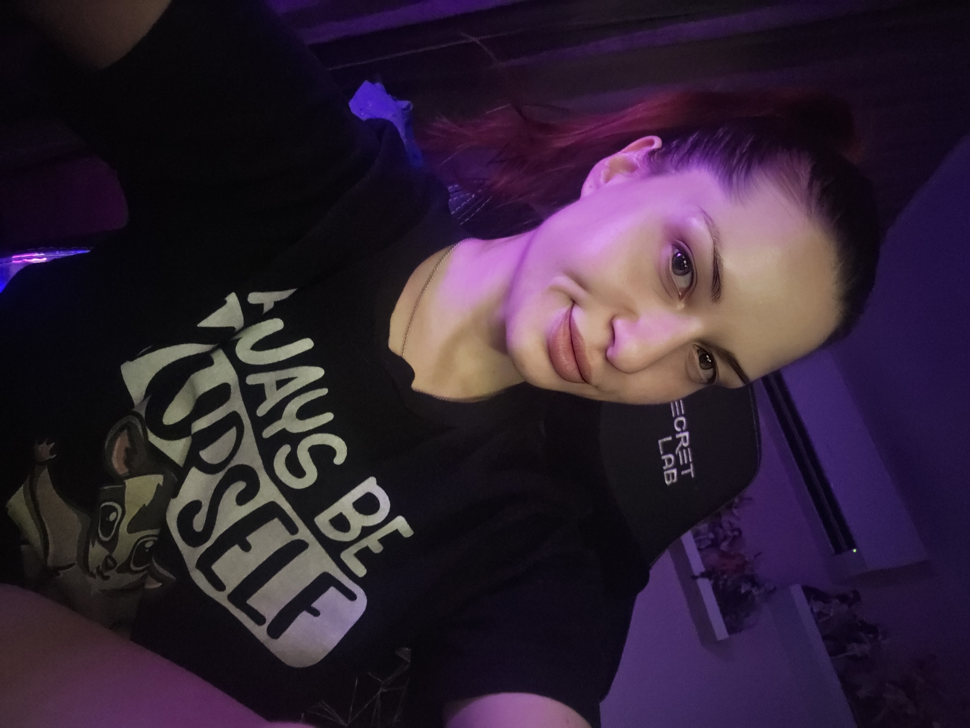 A woman with auburn hair pulled back in a ponytail is taking a selfie in a room lit by purple ambient light. She is wearing a black t-shirt with white text that reads "Always Be Yourself" and features a cartoon animal graphic. Behind her, a black gaming chair with the "Secret Lab" logo is visible, and shelves with figurines are mounted on the purple wall. The overall impression is that of a casual, personal space, likely a bedroom or gaming area. The specific location in Burgas, Bulgaria cannot be determined from the image alone.