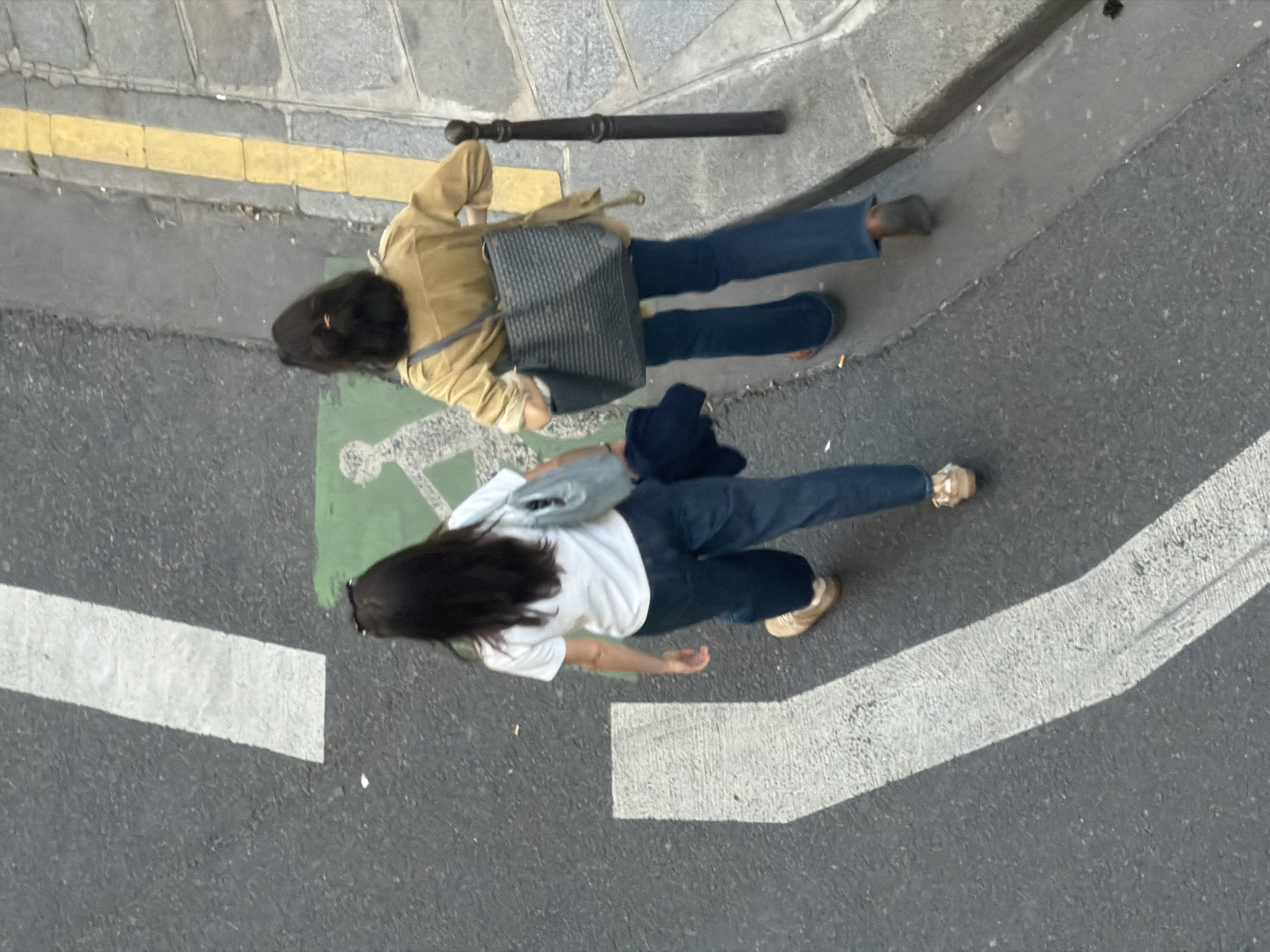 The image shows an aerial view of a section of a street in Paris, France. Two women are walking on the sidewalk and marked crosswalk area of the street. The first woman is wearing a tan jacket, blue jeans, and dark shoes. She has a gray purse slung over her shoulder, and a dark, possibly rolled-up item is on the ground near her. The second woman is wearing a white t-shirt, blue jeans, and light-colored shoes, and she has a light-colored purse over her shoulder. A dark object, possibly a jacket, is held in one arm. The street features typical Parisian elements: stone sidewalk with a yellow line, a curb, and an asphalt street surface with white crosswalk lines and a wheelchair-accessible symbol painted on the ground.