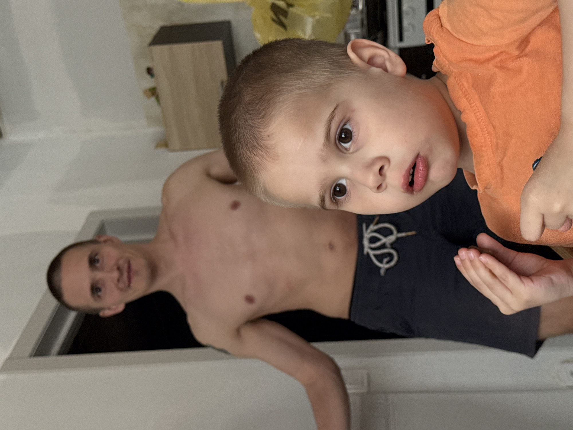 A man and a young child are in a room. The man is shirtless and wearing dark shorts with a design on the front. The child is wearing an orange shirt. The location appears to be a residential building in Chaikovsky, Russia, although this cannot be determined for certain from the image. The child is holding their hand up in front of them.
