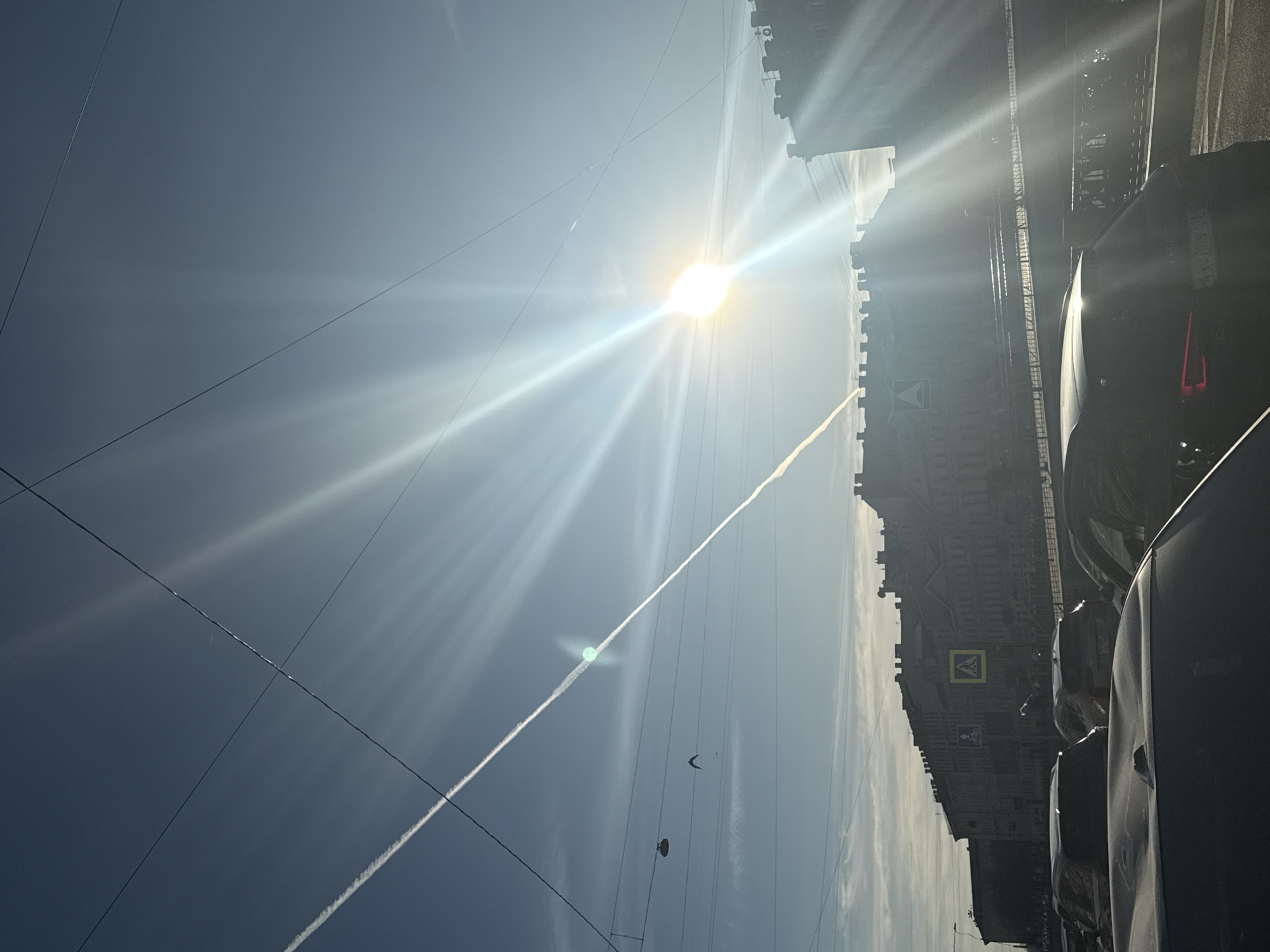 The image shows a street scene in Saint Petersburg, Russia, under a bright sun. The sky is blue with a contrail from an aircraft. Power lines intersect the sky. The right side of the image shows a building facade and parked cars along the side of the street. Road signs are visible on the building.