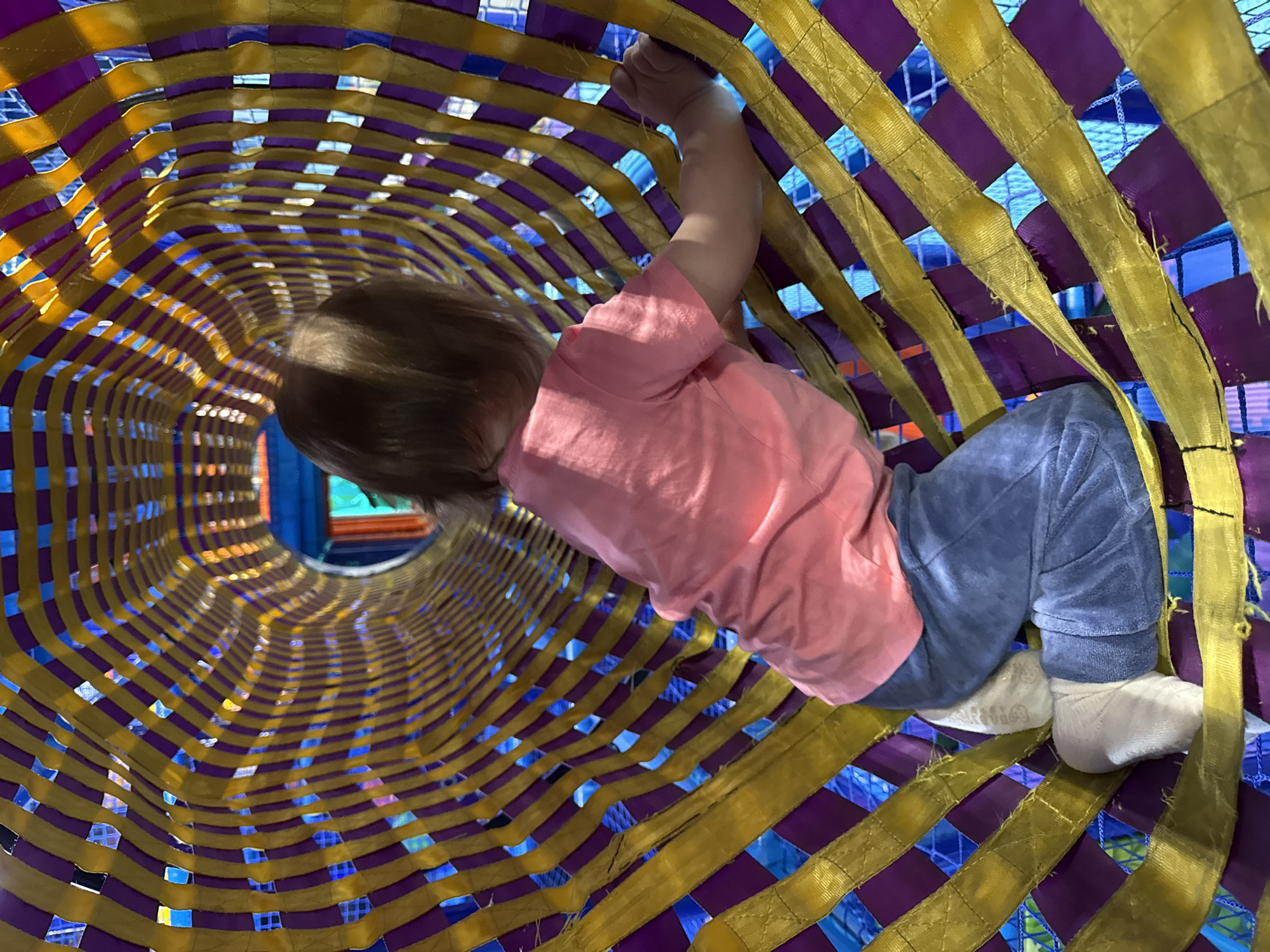A child crawls through a brightly colored tunnel structure. The tunnel is made of purple and yellow bands woven together, allowing glimpses of blue and other colors through the gaps. The child wears a pink shirt and blue pants, with white socks on their feet. Their back is towards the camera as they navigate the tunnel. The location is not discernible as Перовское сельское поселение, Ukraine.