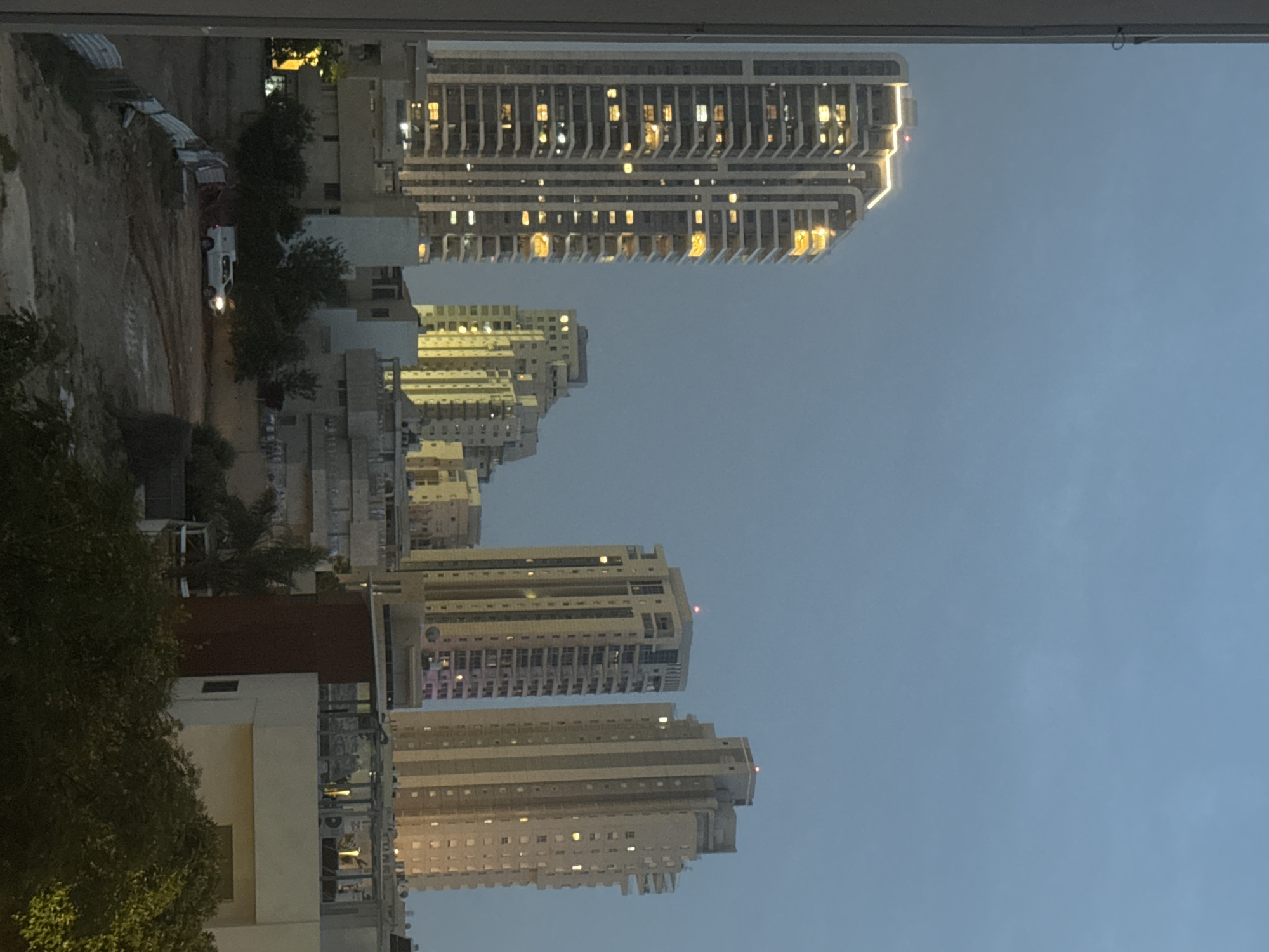 The image shows a night-time view of buildings in Netanya, Israel. The shot appears to be taken from a high vantage point, likely a building or upper floor. Several tall, multi-story buildings dominate the skyline, with lights visible in many windows, indicating residential or commercial use. In the lower left foreground, trees and buildings are visible, suggesting the perspective is looking over a more established part of the city towards newer, taller structures. A vacant lot is also visible. The sky is a dusk or evening color, suggesting a time after sunset.