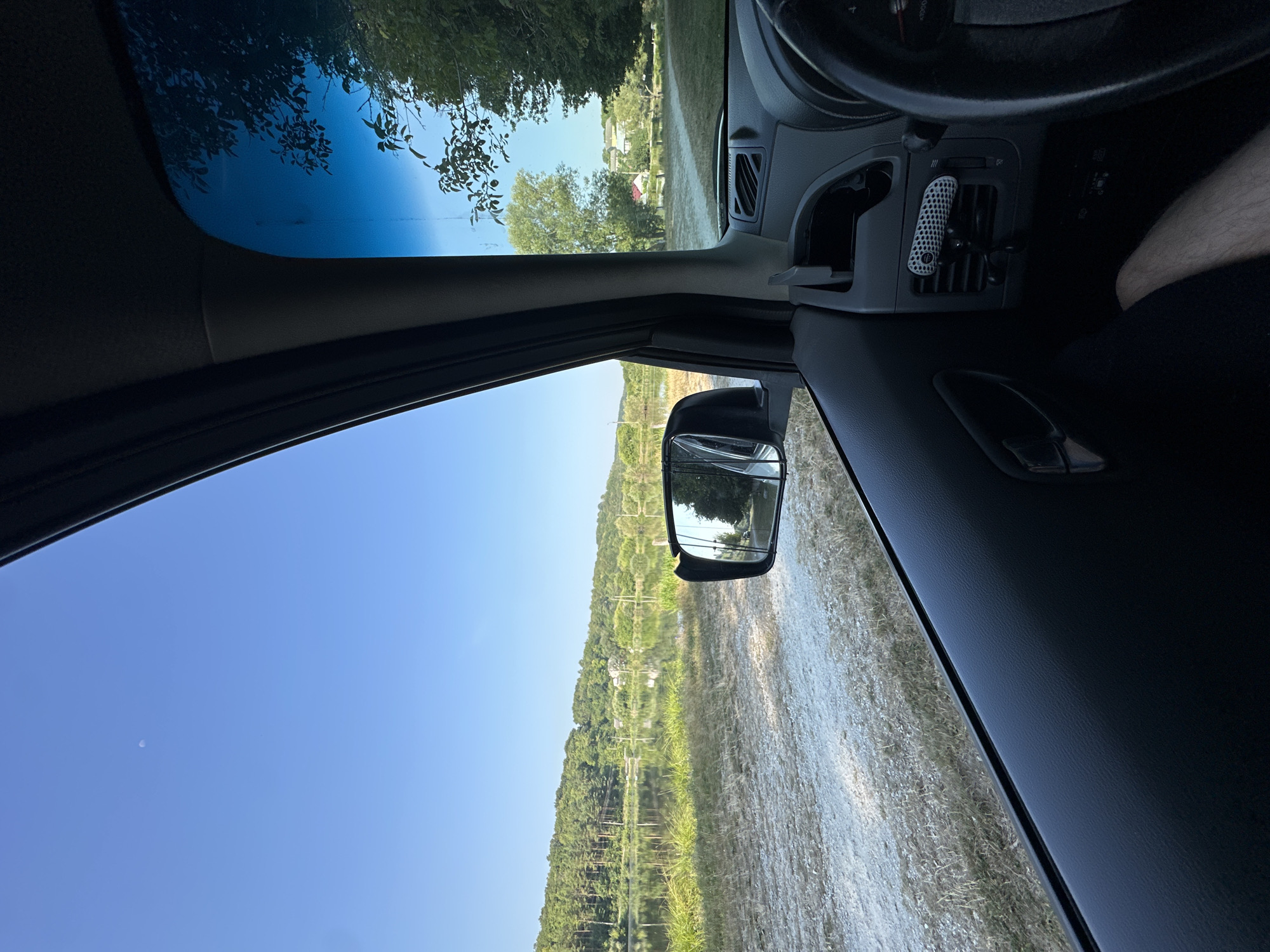 The image is an interior view from the driver's side of a vehicle looking out the window and windshield. Outside, there is a calm body of water reflecting a tree-lined hilly landscape under a clear blue sky. A dirt road runs along the edge of the water. The interior of the car shows the side door panel, a rear-view mirror, and part of the dashboard with a steering wheel and air vent. A small, textured air freshener is clipped to the air vent. A person's leg and knee are visible in the lower right of the frame. The location is identified as Makiv, Ukraine, and the scene depicts a peaceful, rural setting by the water.