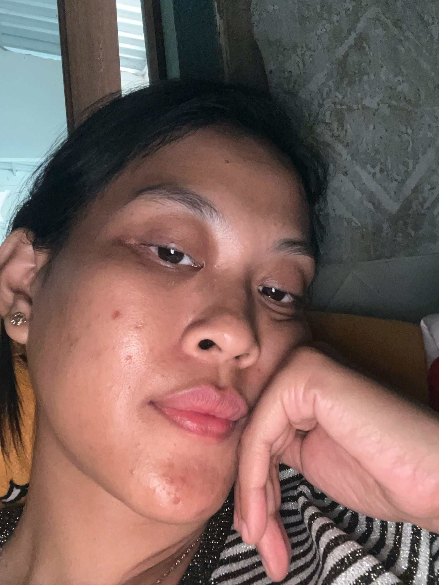 A close-up selfie shows a person's face, with their left hand resting on their chin and cheek. The background is dark and textured, appearing to be a wall. The person's hair is dark and pulled back. They are wearing a striped shirt. The setting is indoors, likely a home, and based on the metadata, is located in Kecamatan Pati, Indonesia. The lighting is dim, creating shadows. The person's gaze is directed towards the camera.