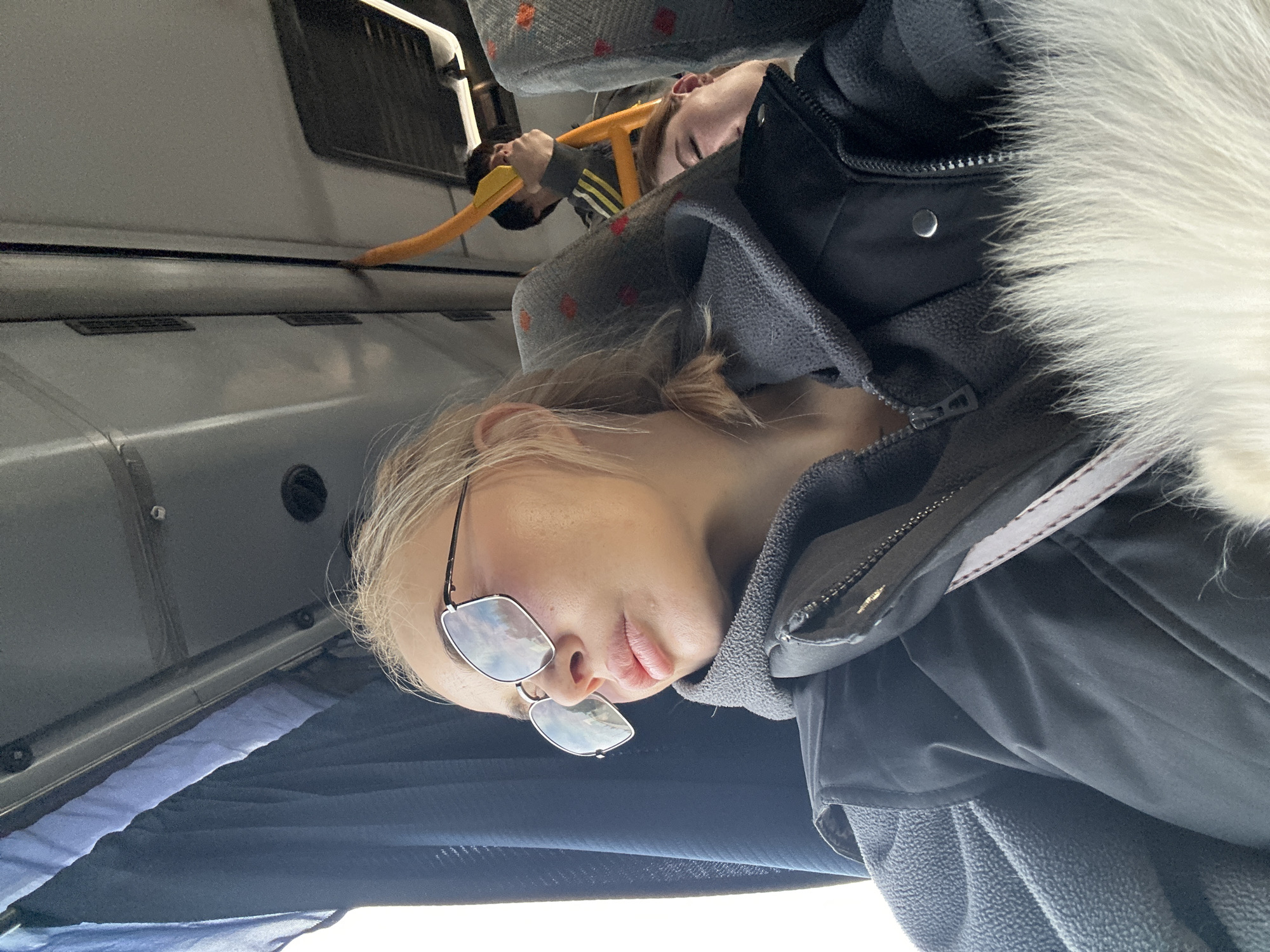 A bus interior is shown in a photo. The foreground is dominated by a woman with blond hair and sunglasses wearing a winter jacket with fur trim. She appears to be lying down, possibly sleeping, as the camera angle is from above. Behind her, another person is holding onto a yellow overhead bar, also seemingly reclining or sleeping. Bus seats with a patterned fabric cover are visible. Overhead storage or compartments run along the side of the bus. The location, Megion, Russia, cannot be definitively confirmed from visual elements alone.
