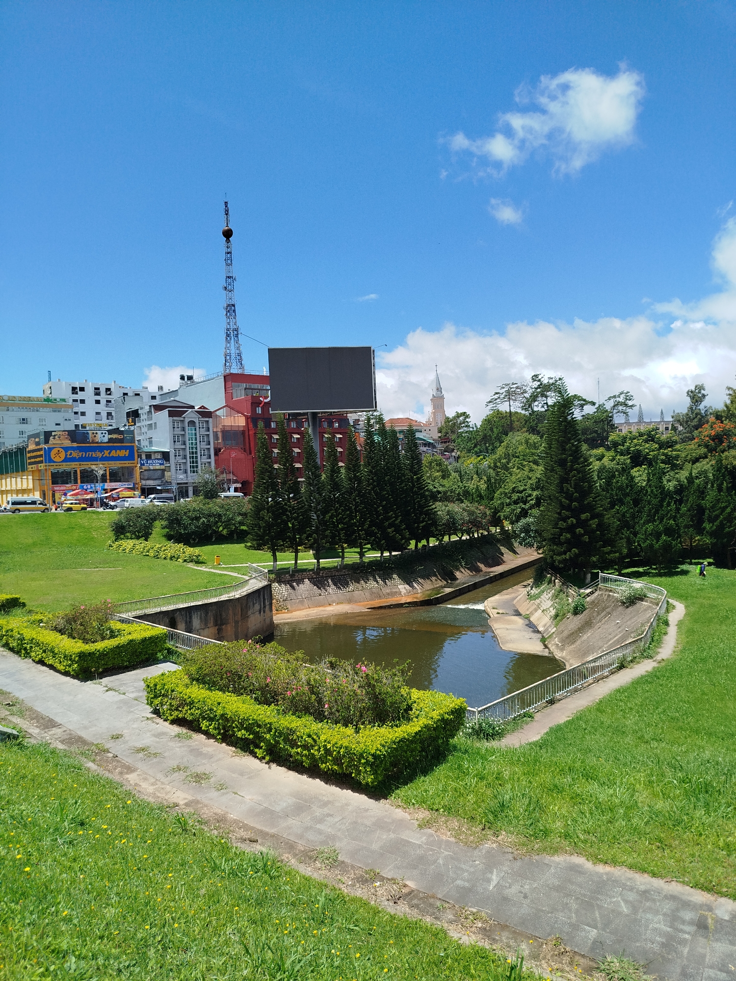 The image shows a park area in Da Lat, Vietnam, on a clear day with scattered clouds. In the foreground, a paved path winds through a grassy slope dotted with small yellow wildflowers. To the left, meticulously manicured hedges form two rectangular beds. The path leads to a canal with concrete embankments.
Beyond the canal, a row of tall, slender pine trees stands out. Further in the background, a cityscape is visible, featuring various buildings of different heights and architectural styles. Notably, a tall telecommunications tower with a spherical object at its apex dominates the skyline. A large, dark rectangular billboard is mounted on a building to the left of the tower. A church spire with a pale, cream-colored facade is visible above the trees on the right side of the image, suggesting a location near a religious institution. The overall environment is a blend of urban elements and green spaces, characteristic of Da Lat's landscape. There are no people clearly visible in the scene.