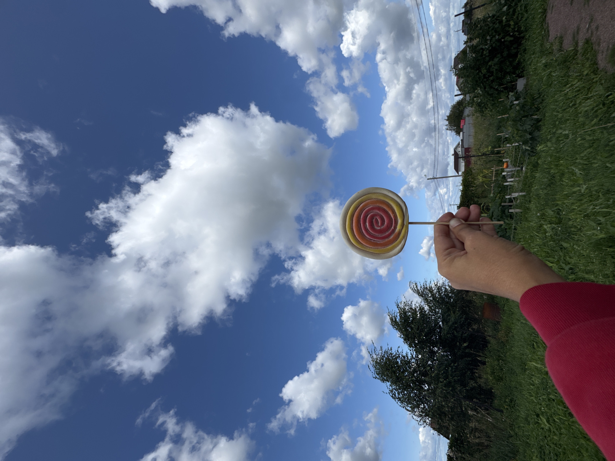 A hand in a red sleeve holds a colorful, swirled lollipop against a bright blue sky with scattered white clouds. The background depicts a rural landscape with green grass, trees, and several houses. The location is identified as Kempelevo, Russia.
