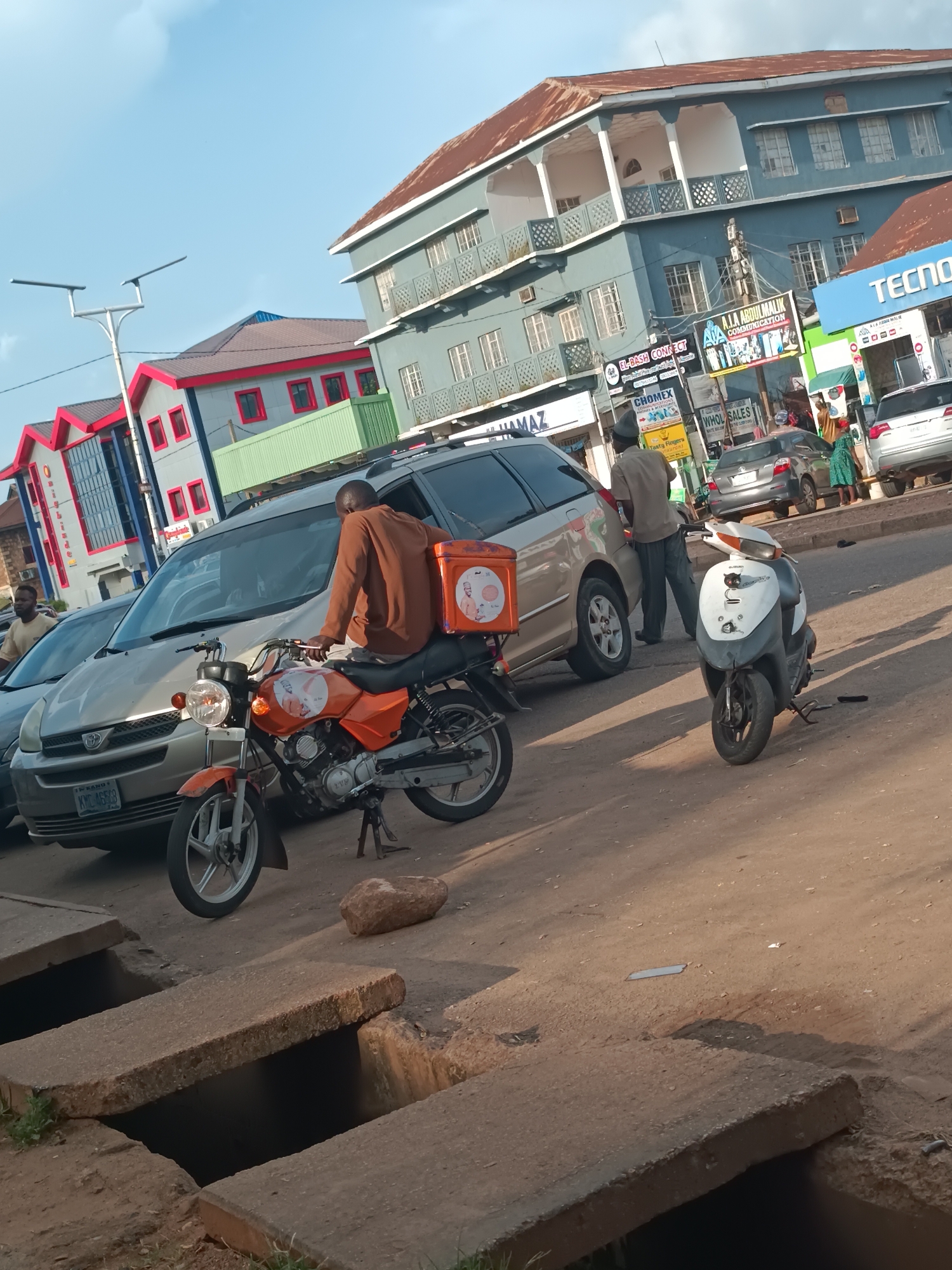 The image depicts a street scene in Jos, Nigeria, characterized by a mix of residential and commercial buildings under a partly cloudy sky. In the foreground, a man is seated on an orange motorcycle, facing towards the right. The motorcycle has a delivery box mounted on its rear, adorned with an image of what appears to be a person in uniform. The man is wearing a brown long-sleeved shirt and dark pants. Next to the motorcycle is a light-colored car. Further down the street, another individual stands near a silver minivan. Various signs are visible on the buildings, including "TECNO," "CHOMEX," and "A.A. ABDULMALIK COMMUNICATION," suggesting a commercial area. The road surface is paved, and concrete drainage covers are present along the roadside. The overall impression is of an urban environment during daylight, with activity suggesting daily life and commerce.