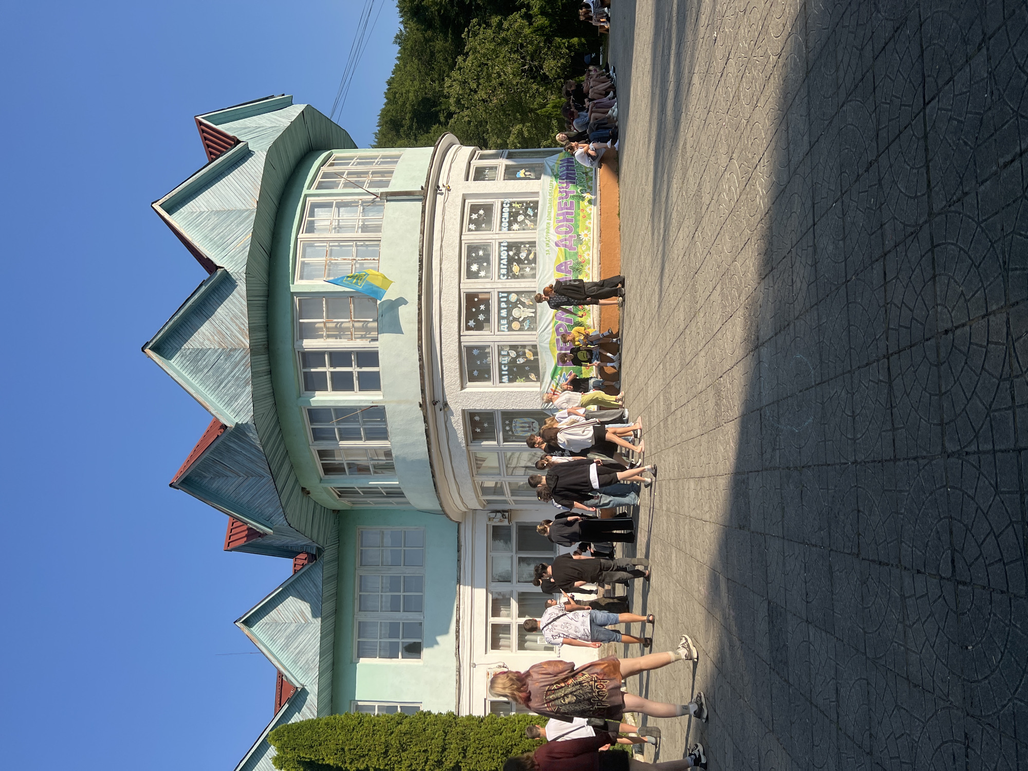 The image shows a sunny outdoor scene in front of a light teal building with a distinctive red-roofed, multi-peaked architectural design. A Ukrainian flag is visible on the building's upper level. A group of people, predominantly young adults and teenagers, are gathered on a paved courtyard. Some are standing and talking, while others are seated on a low wall or the ground. A banner with text and floral imagery is displayed on the lower part of the building. The ground is paved with square tiles, some of which have circular patterned inlays. A tall green conifer tree stands to the left of the building, and trees line the background. The scene appears to be a public space, possibly near a school or community center in the village of Сонячне, Ukraine.