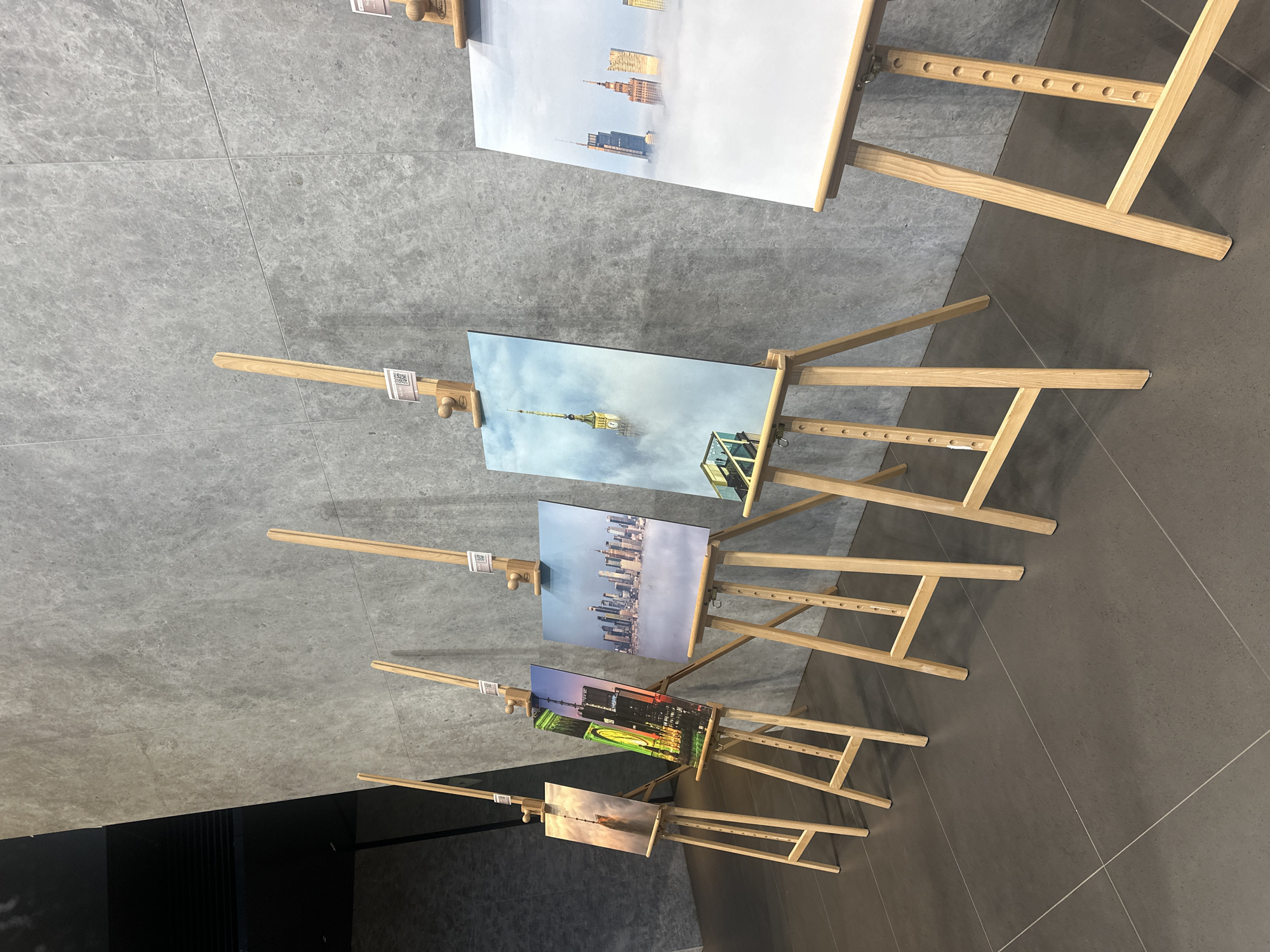 The image shows a row of five wooden easels, each displaying a photograph of a cityscape. The photographs depict skyscrapers, some emerging from clouds, creating a surreal and dramatic effect. The easels are arranged in a gallery-like setting against a textured, grey wall. The floor is tiled with large, dark grey tiles. The lighting appears to be artificial, casting soft shadows. The artworks are positioned in a line, suggesting an exhibition. One of the artworks features the Palace of Culture and Science in Warsaw, Poland, identifiable by its distinctive architecture and spire, surrounded by mist. The remaining artworks display similar architectural styles and atmospheric conditions, potentially from different cities or with artistic interpretations. There are no people visible in the scene.
