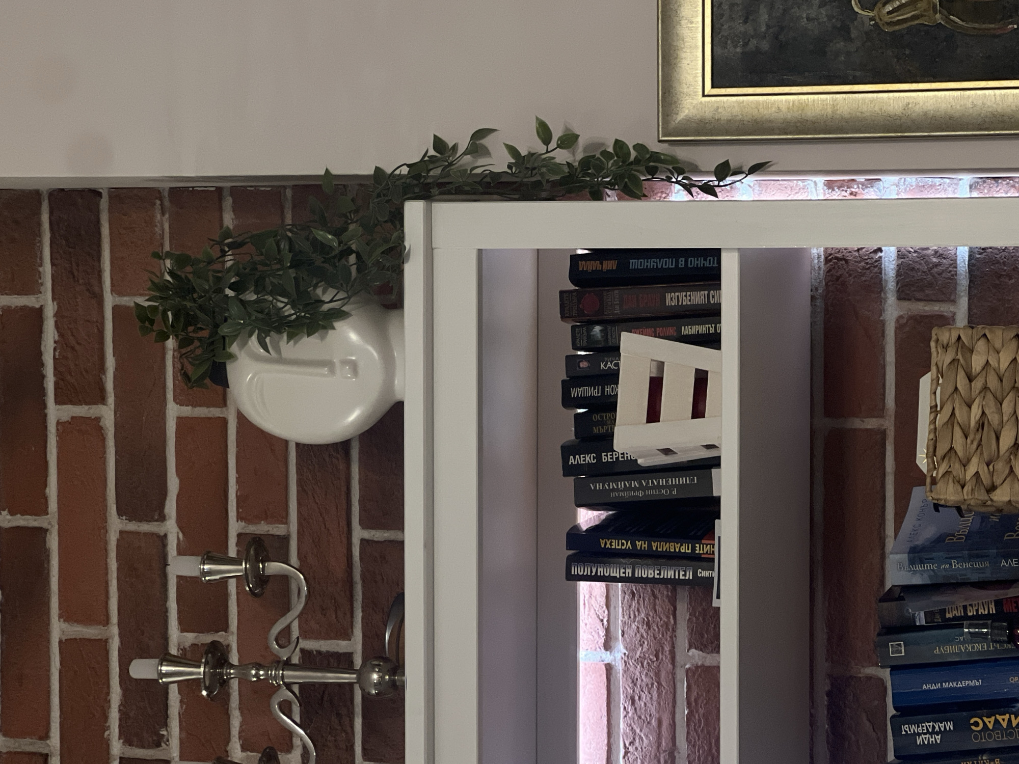 The image shows a section of an interior brick wall adorned with decorative elements. On the left, a white vase with a plant attached to the wall is visible. A silver candelabra hangs below the vase. To the right, a white bookcase is filled with books, some of which have titles in Cyrillic. The bookcase obscures a view of the brick wall behind it. The top of a framed picture is visible in the upper right corner. There is no information in the photo to determine if this is in Sofia, Bulgaria.