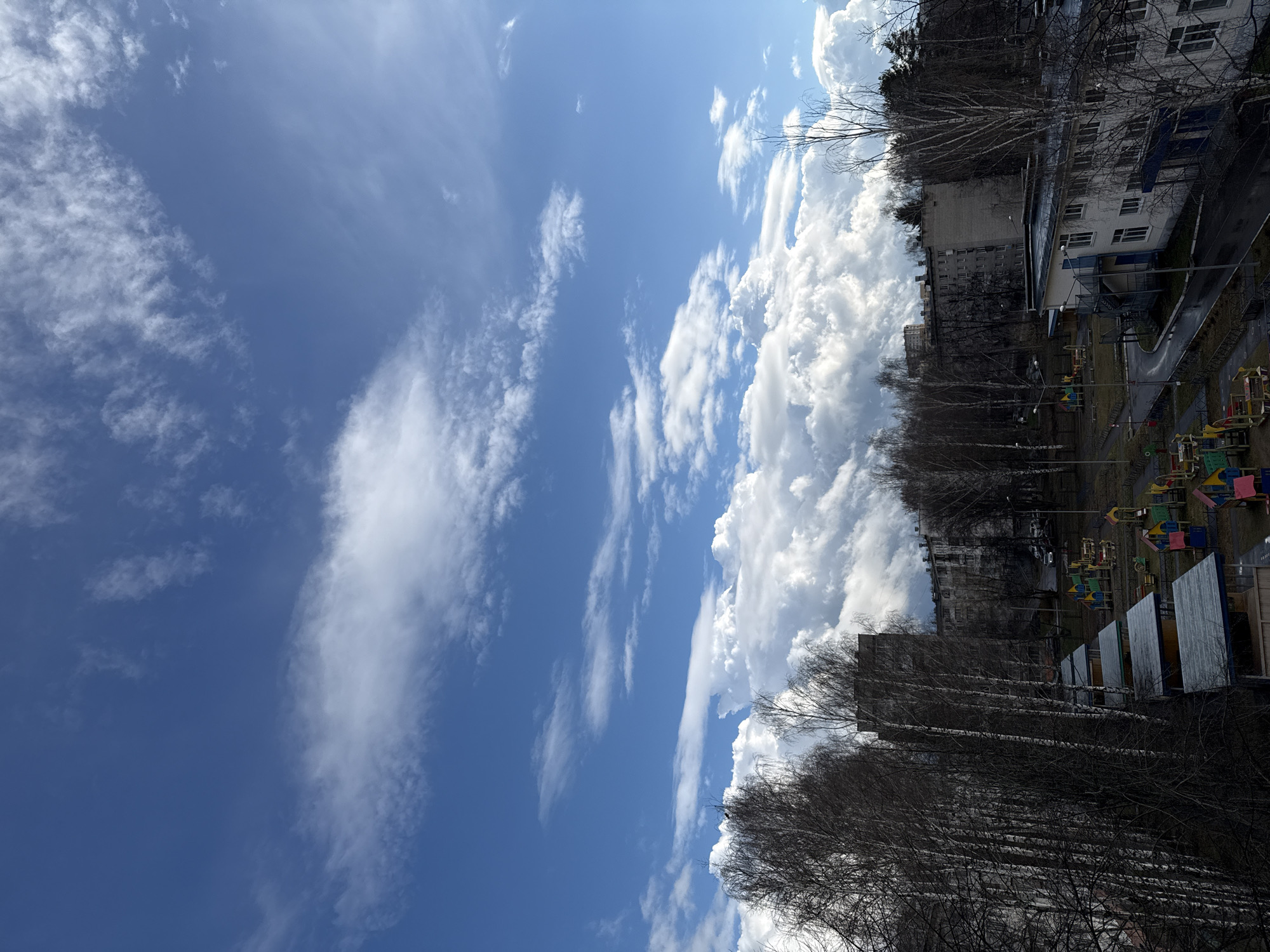 The image presents a view in Chaikovsky, Russia, featuring a juxtaposition of natural elements and urban structures. The sky occupies a significant portion of the frame, displaying a gradient from deep blue to lighter shades with streaks of cirrus clouds and large cumulonimbus formations. On the right side of the image, there are multi-story residential buildings, bare trees, and a playground. The playground features colorful structures. The scene appears to be viewed from an elevated position, possibly from another building or higher ground. The lighting suggests it is daytime.