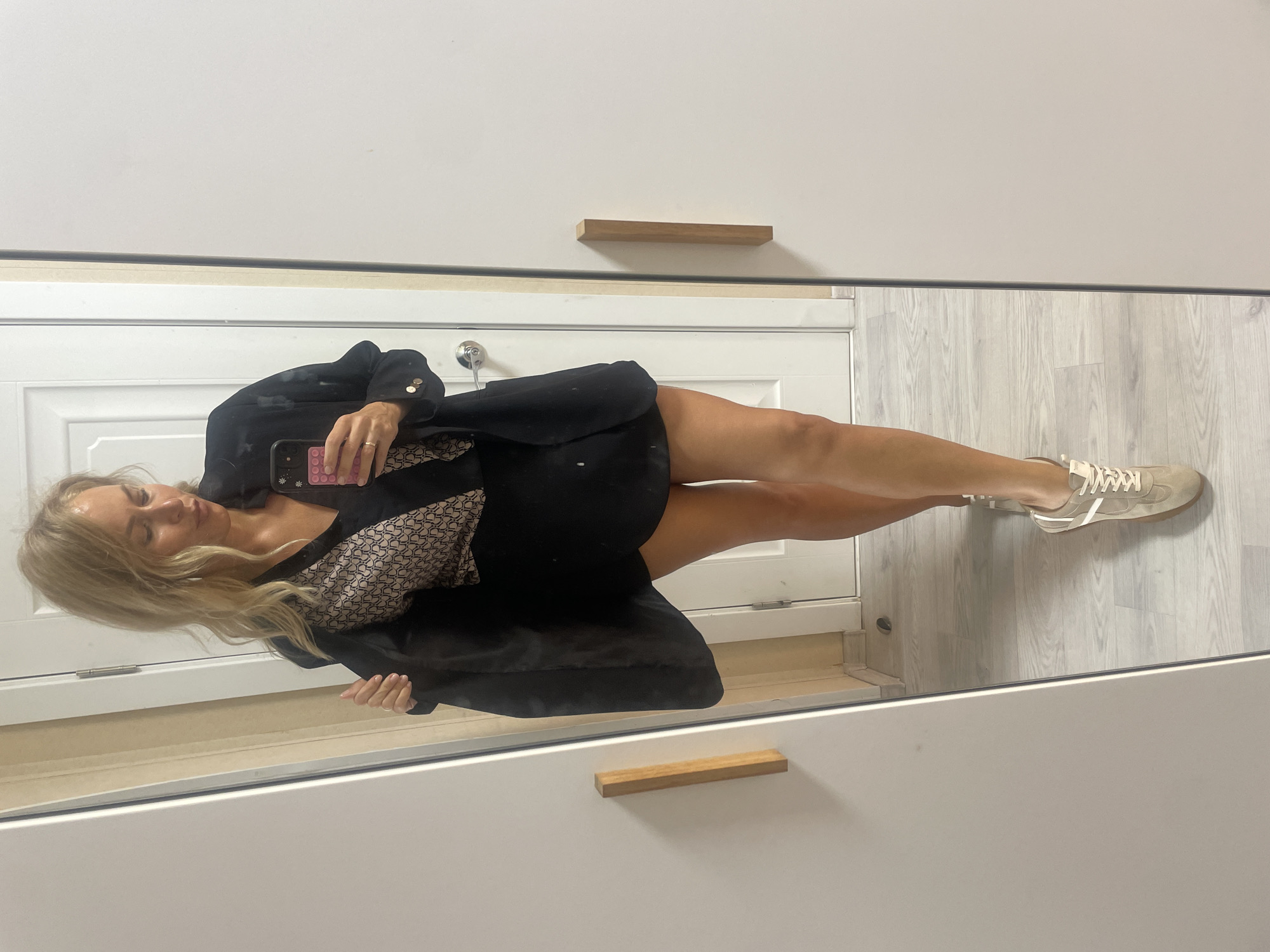 A blonde woman in a black blazer and patterned top is taking a mirror selfie. She is holding a phone with a pink and black case. Her legs are visible and she is wearing beige sneakers. The background appears to be a bathroom or wardrobe area, featuring a white door and cabinets with wooden handles. The floor is a light grey wood-patterned surface. The location appears to be indoors. There is no indication this image was taken in Baranovka, Russia.