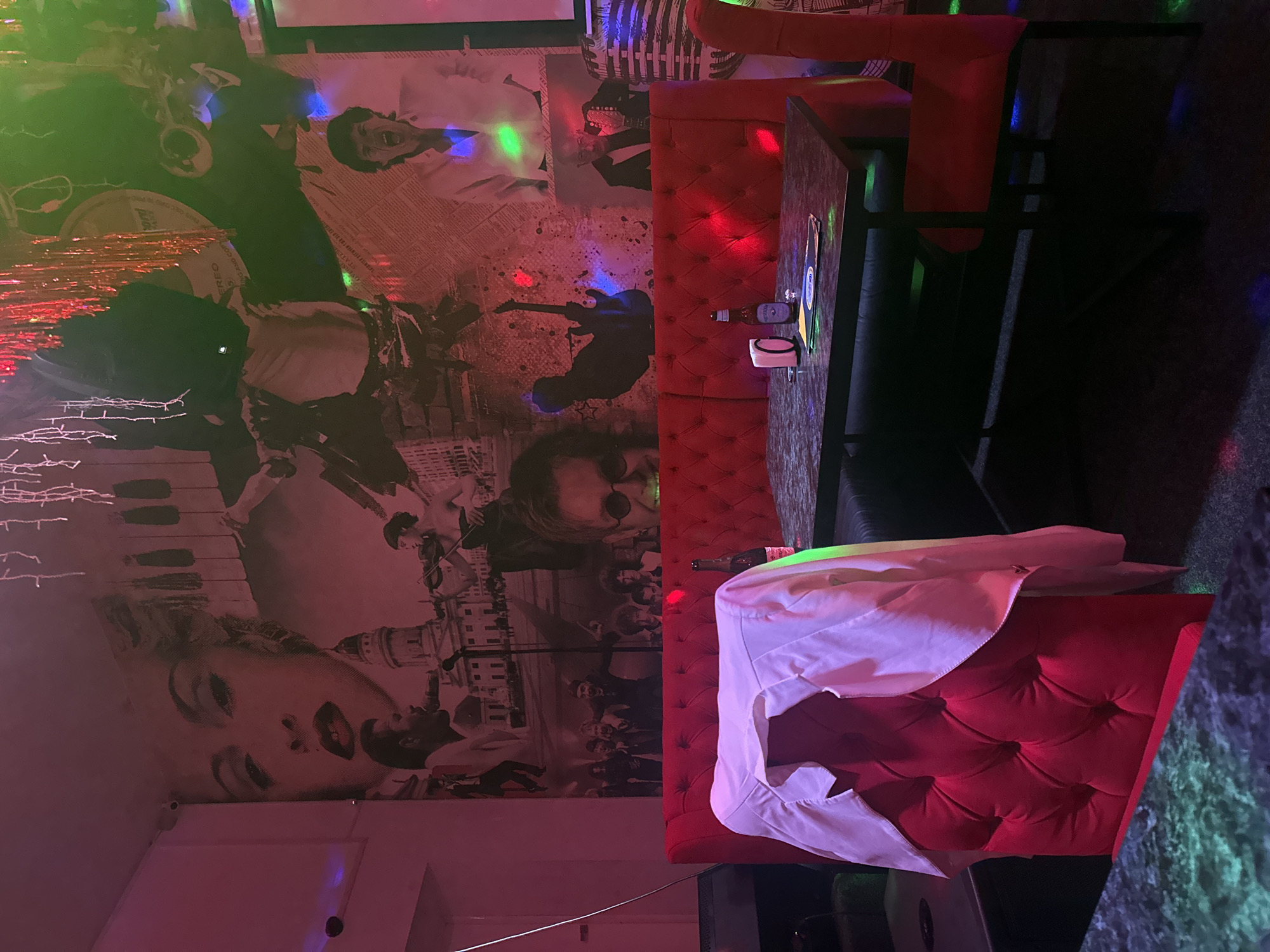 This image depicts the interior of a dimly lit establishment, possibly a bar or club, with red upholstered seating and a table. The walls are adorned with a large mural featuring stylized portraits and scenes, including what appears to be a singer, a violinist, and possibly musicians. The lighting is dramatic, with colored spotlights and decorative string lights, creating a vibrant and somewhat retro atmosphere. A white shirt is draped over the back of a red chair in the foreground. On the table, there are two bottles, one partially visible, and a small white object, possibly a napkin dispenser. The overall impression is of a lively entertainment venue. There is no visible indication of the city of Chaikovsky, Russia within the image.