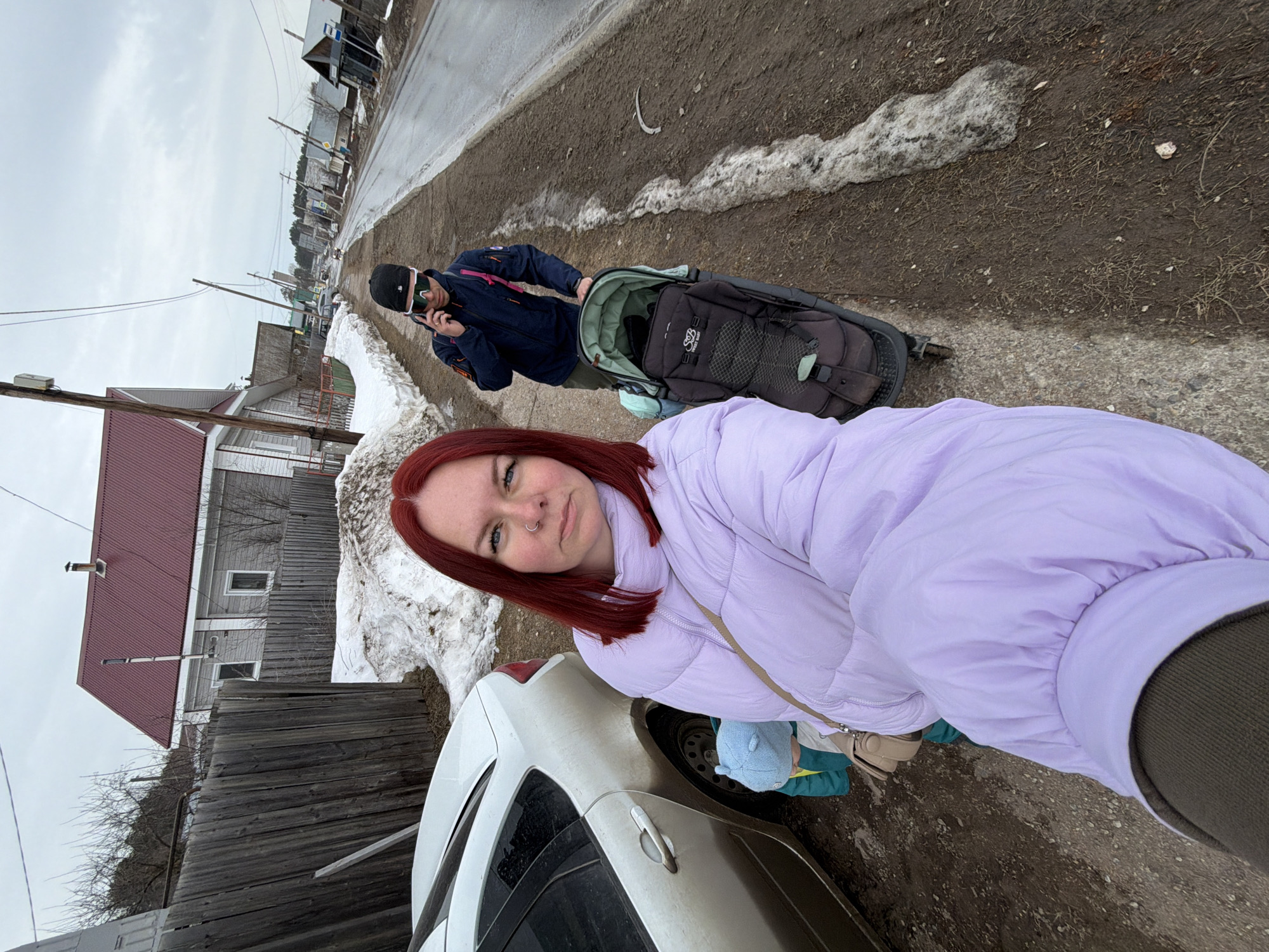 This image depicts an outdoor scene on a street in Chaikovsky, Russia, characterized by muddy ground and melting snow under an overcast sky. In the immediate foreground, a woman with red hair and light blue eyes, wearing a lilac puffer jacket and a beige cross-body bag, is capturing a selfie, looking directly into the camera. She has a ring piercing on her left nostril. To her left, partially obscured by a dirty white sedan and the woman herself, a small child is visible, wearing a light blue hat and a teal jacket. Further back, on a muddy dirt shoulder adjacent to a wet, paved road, a man is walking. He is wearing a dark blue jacket, a black cap, and wrap-around sunglasses, and is holding a smartphone to his right ear, engaged in a call. He is pushing a dark-colored baby stroller with a light green interior, displaying the "SWEET BABY" brand. The background features a residential area with several wooden houses, some with light-colored siding and red corrugated metal roofs, partially hidden behind a weathered wooden fence. Wooden utility poles with overhead wires line the road, which recedes into the distance. Dirty piles of melting snow are scattered along the roadside and adjacent to the fence.
