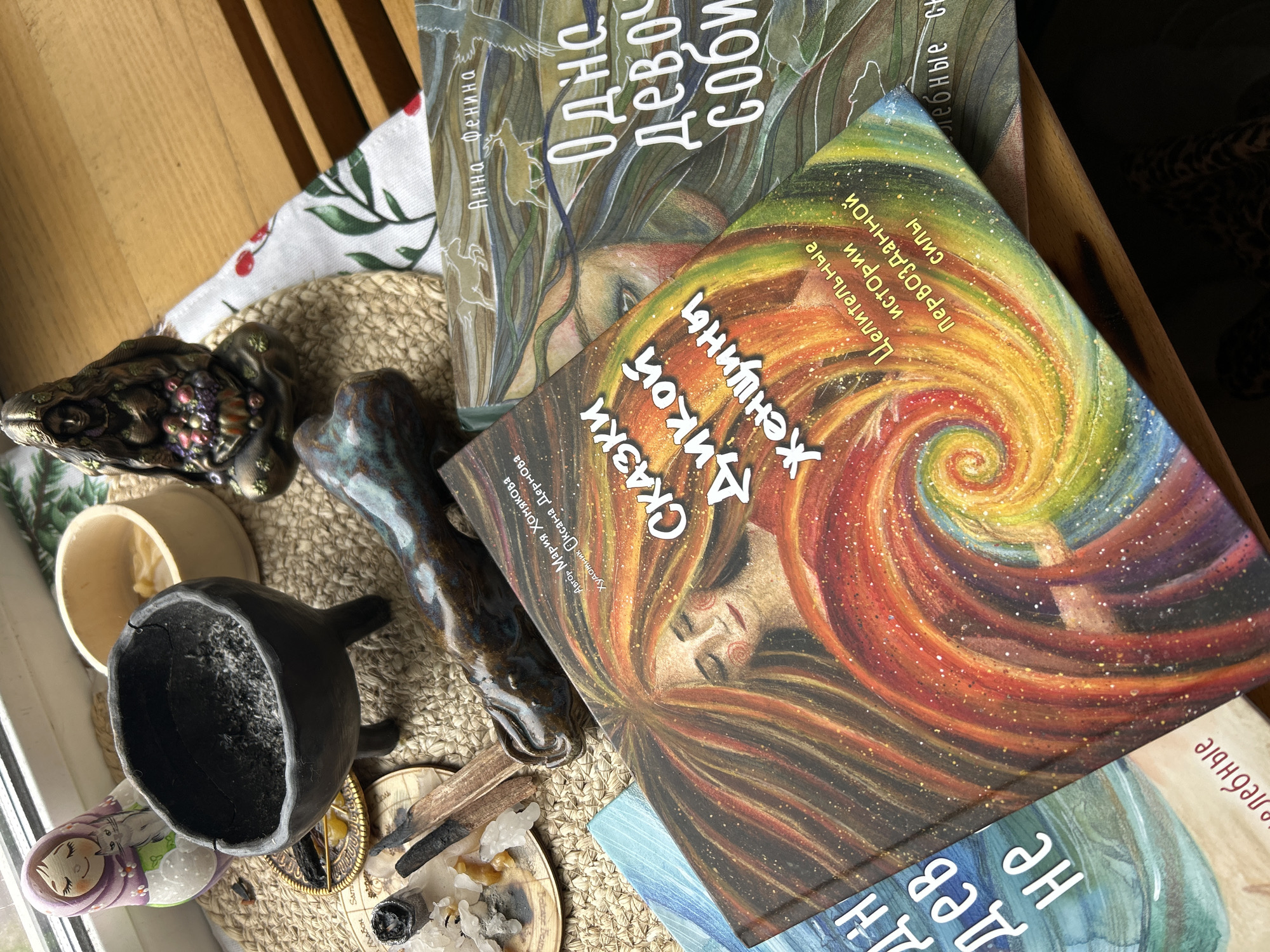 The image is a close-up shot taken indoors, displaying a collection of objects arranged on a tabletop, possibly near a window.
The main focus of the image are two books with Russian text. The book titles in Russian are "Сказки Дикой Женщины" and "Одна девочка соби".
A small black cauldron filled with what looks like ashes or dark material is in the lower left corner. Next to it are what appear to be ritual items: a small wooden bowl with a light-colored substance inside, a patterned ceramic object, a blue-glazed ceramic figurine, and a small dish containing crystals and burnt incense or herbs. A Russian nesting doll (Matryoshka doll) is partially visible beside the cauldron.
The objects are resting on a woven mat and a white cloth with a red and green cherry and leaf pattern. Part of a wooden chair can be seen in the background, with a blurred pattern visible through an opening in the chair.
The lighting suggests natural light coming from a window, casting shadows and highlights on the objects. The scene has a mystical or spiritual feel due to the books and the ritualistic objects.
There is no indication in the image that indicates the location of the city of Moscow.