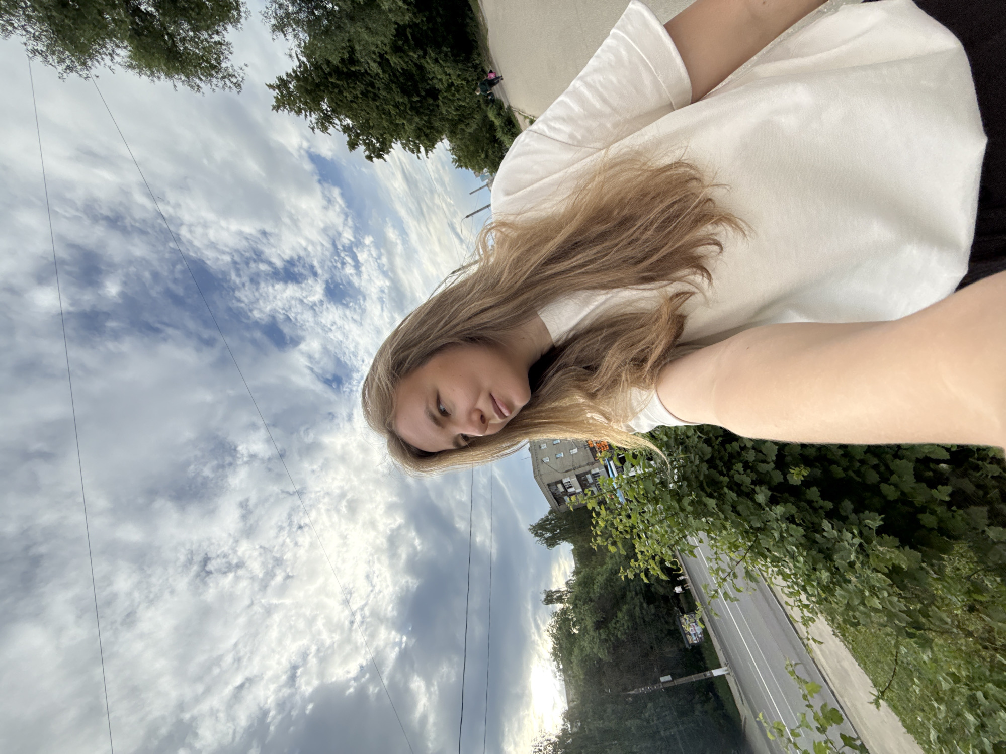A woman with long, blonde hair is taking a selfie on a cloudy day. She is wearing a white t-shirt and black pants. She is standing on a sidewalk next to a road with trees and buildings in the background. There are power lines visible in the sky. The location is the city of Vladimir, Russia.