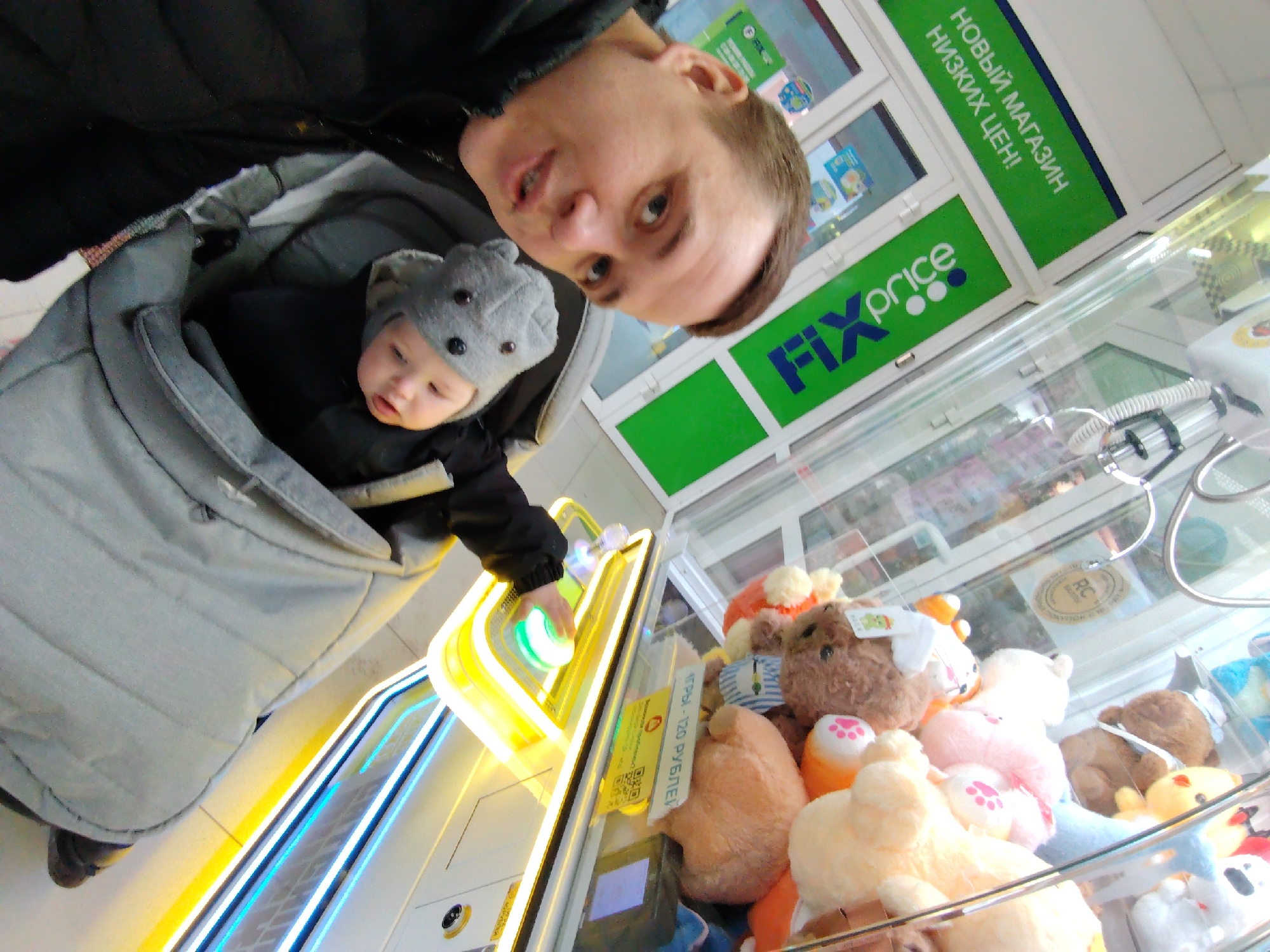 A low-angle shot of an adult and a baby, likely inside the "Fix Price" store, in Chaikovsky, Russia. The adult stands next to a claw machine filled with plush toys. The baby, wearing a grey hat with ears, is in a carrier worn by the adult. The baby's hand is reaching out to press the green button on the claw machine. Above the machine is a sign in Russian "NEW STORE LOW PRICES!".