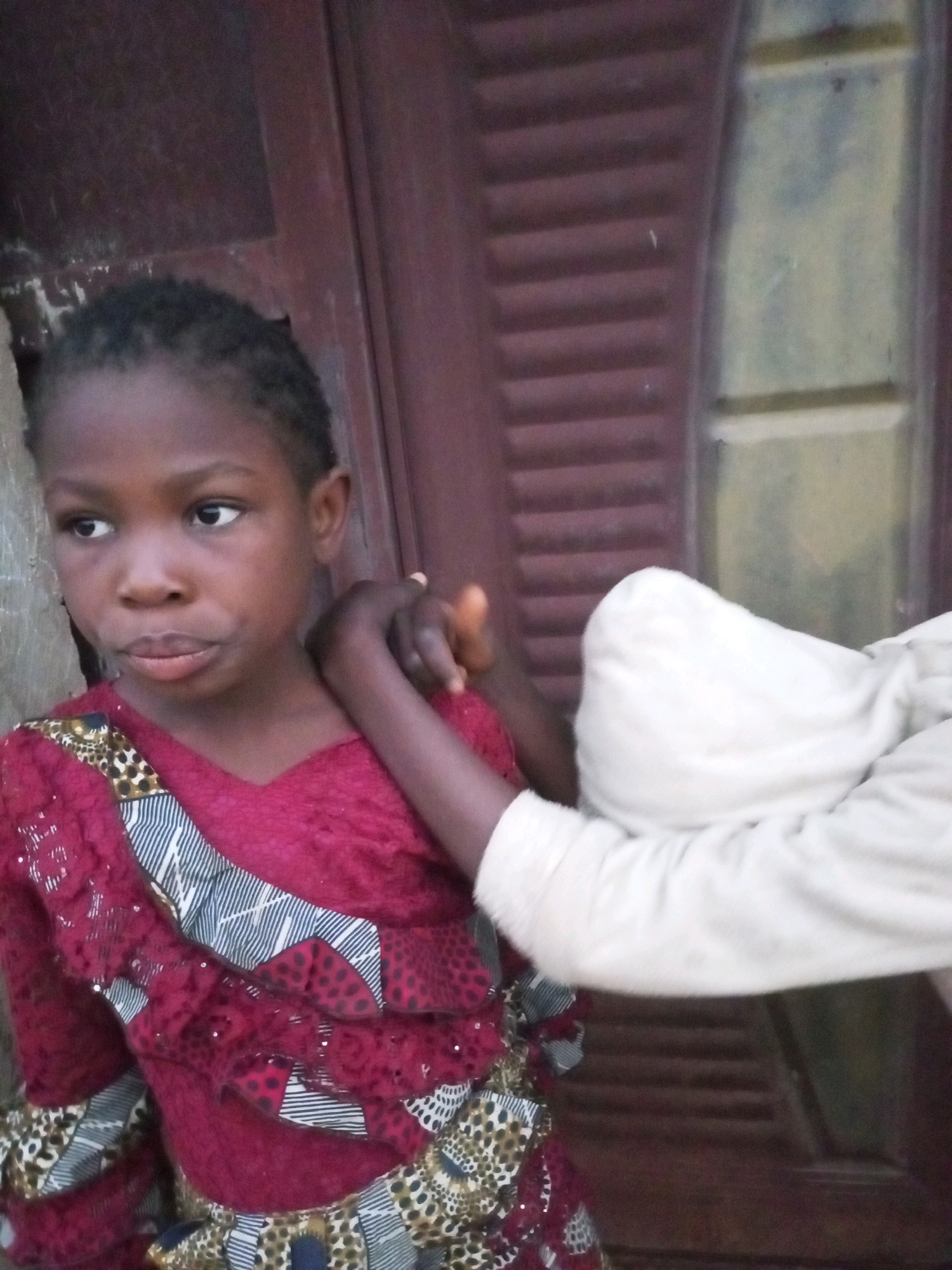 A close-up, eye-level, medium shot captures a young Black girl in the foreground, looking to her left. Her head is slightly tilted, and her mouth is gently pursed. She has dark, short hair and wears a maroon, textured top with a decorative trim of black and white stripes, red polka dots, and leopard print. Her left arm is bent, with another hand, presumed to be from another person, resting on her shoulder. The background features a weathered, dark brown door with horizontal slats and a lighter, textured surface to the right, possibly a window or another wall. The overall lighting suggests an indoor or shaded outdoor environment. The image appears to be taken in Tempure, Nigeria.