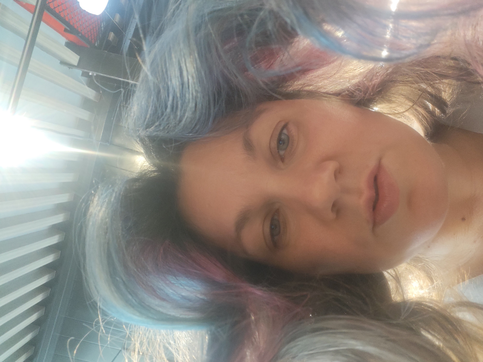 A close-up, tilted selfie depicts a woman with light skin and dyed hair. Her hair has a mix of colors, including blue, grey, and pink. She has light-colored eyes and light pink lips. The background includes a metallic structure of a building. Due to the strong glare of sunlight, the details are not clearly discernible, and no context is provided that would indicate this image was taken in Sholokhovo, Russia.