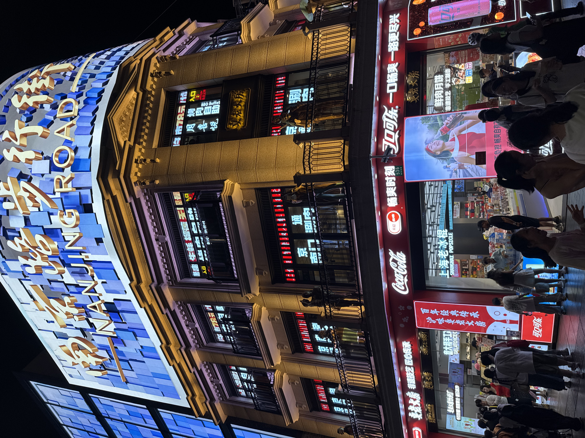 The image shows a nighttime view of Nanjing Road Pedestrian Street in Shanghai, China. The street is illuminated by neon signs and digital displays on the building facades. A large, curved digital billboard at the top of a building displays the words "NANJING ROAD" in golden Chinese characters and English. Below this, the building has multiple floors with balconies, where people can be seen standing. Various businesses are visible at street level, including a Coca-Cola store. People are walking along the street, some are looking at their phones, and others are gathered in front of shops. The overall scene depicts a busy urban commercial area at night.