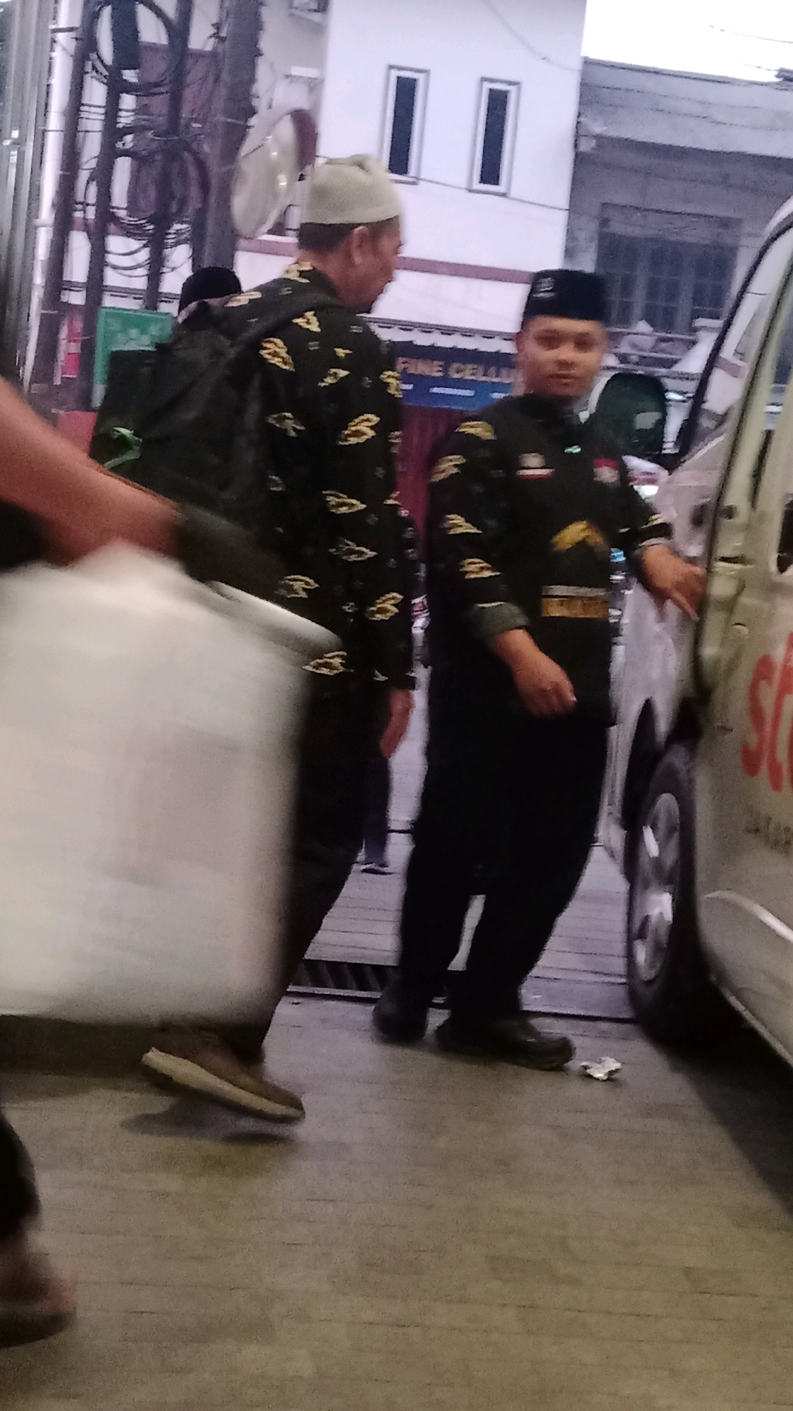 The image depicts a street scene in Tangerang, Indonesia. Two men in dark, patterned shirts and pants, both wearing skullcaps, are present. The man on the left is wearing a light-colored skullcap and the man on the right is wearing a black skullcap. They appear to be standing near a white vehicle, possibly a van, with a portion of its side visible. The man on the right has his right hand resting on the vehicle. A blurry, white object, possibly a bag or container, is in the foreground on the left side of the image, partially obscuring the legs of a person. The ground appears to be paved. In the background, a building with white walls and dark windows can be seen, along with some electrical wires and poles. The lighting suggests it is daytime.