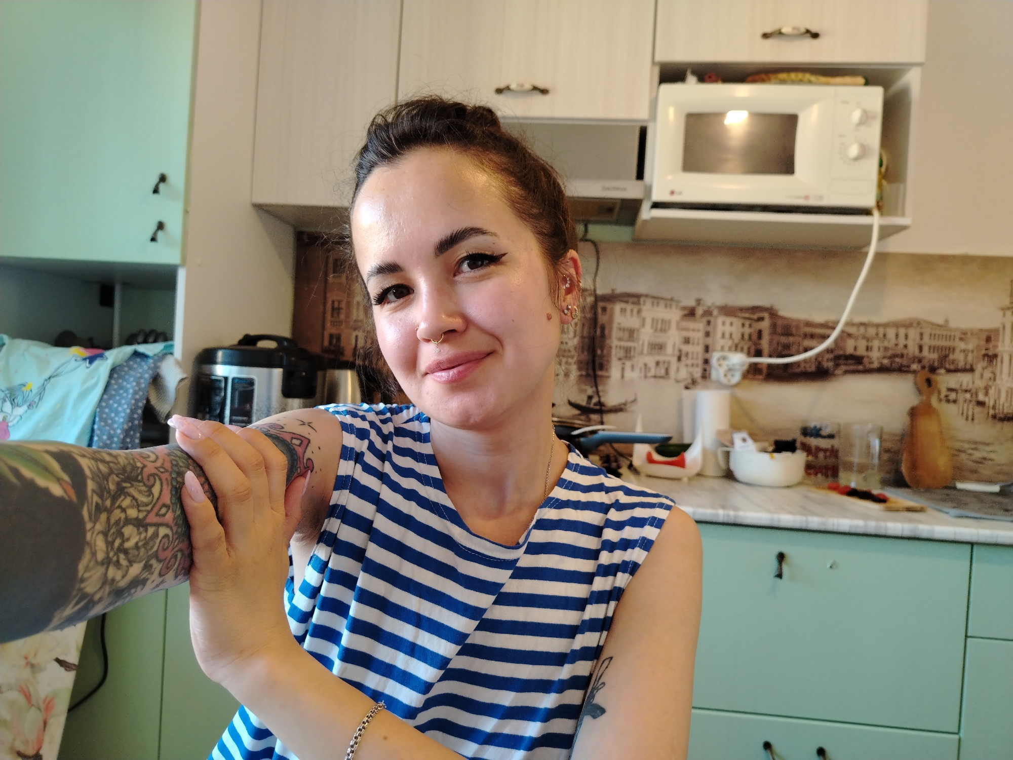 The image depicts a woman in a kitchen. She is wearing a blue and white striped sleeveless top and has tattoos on her arms. Her hair is pulled back in a bun, and she has piercings in her ear and nose. The kitchen has light-colored cabinets, a white microwave, and a backsplash with a picture of a city, possibly Venice. There are various kitchen utensils and appliances visible on the counter. The location is Noviy, Russia.