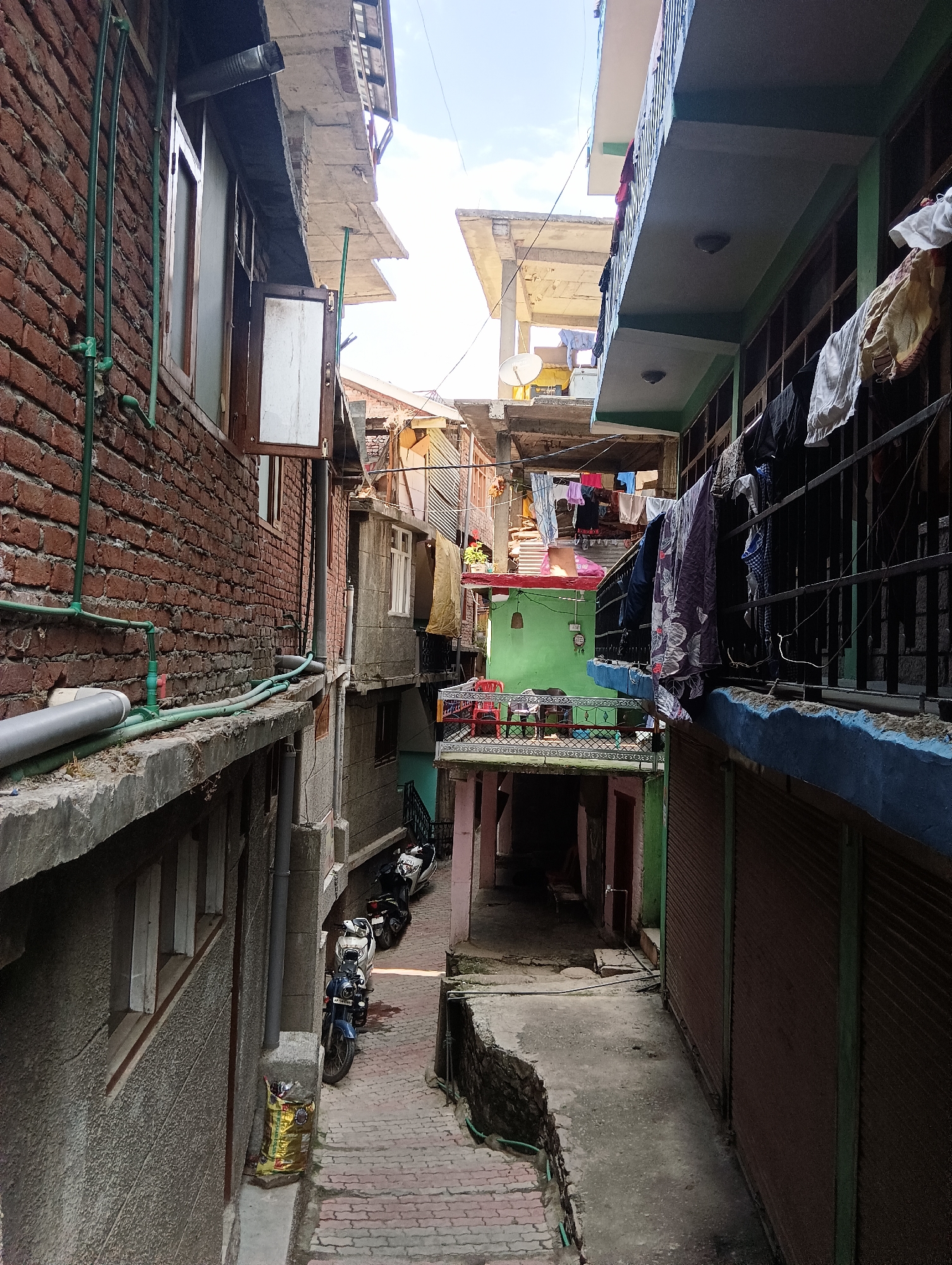 The image depicts a narrow alleyway in a densely built urban area. The buildings on either side are multi-story structures with exposed brick and concrete facades. Several balconies are visible, with laundry hanging to dry on railings. The alleyway is paved with red bricks and is lined with parked motorcycles. One building has a bright green painted facade, and another has a corrugated metal shutter. The sky is visible in the upper portion of the image, suggesting it is daytime. There are no people visible in the scene. The scene appears to be a residential or mixed-use area.